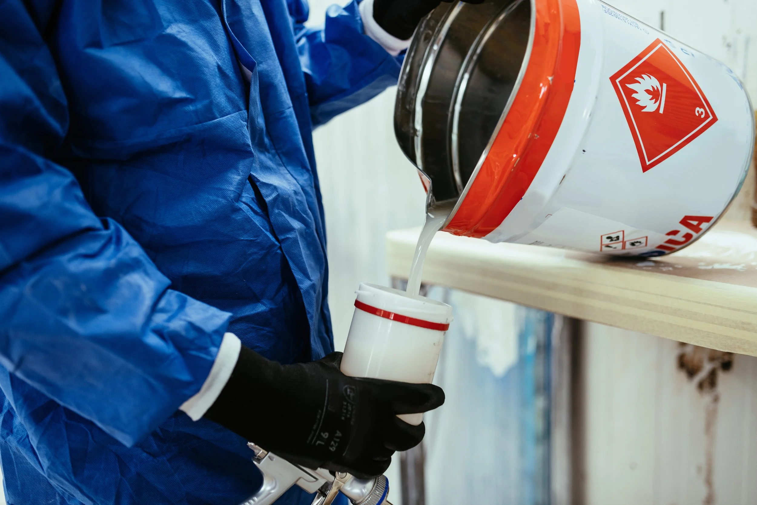 Person wearing blue protective clothing and black gloves pouring a liquid from a container with hazard labels into a white cup in a laboratory or industrial setting.