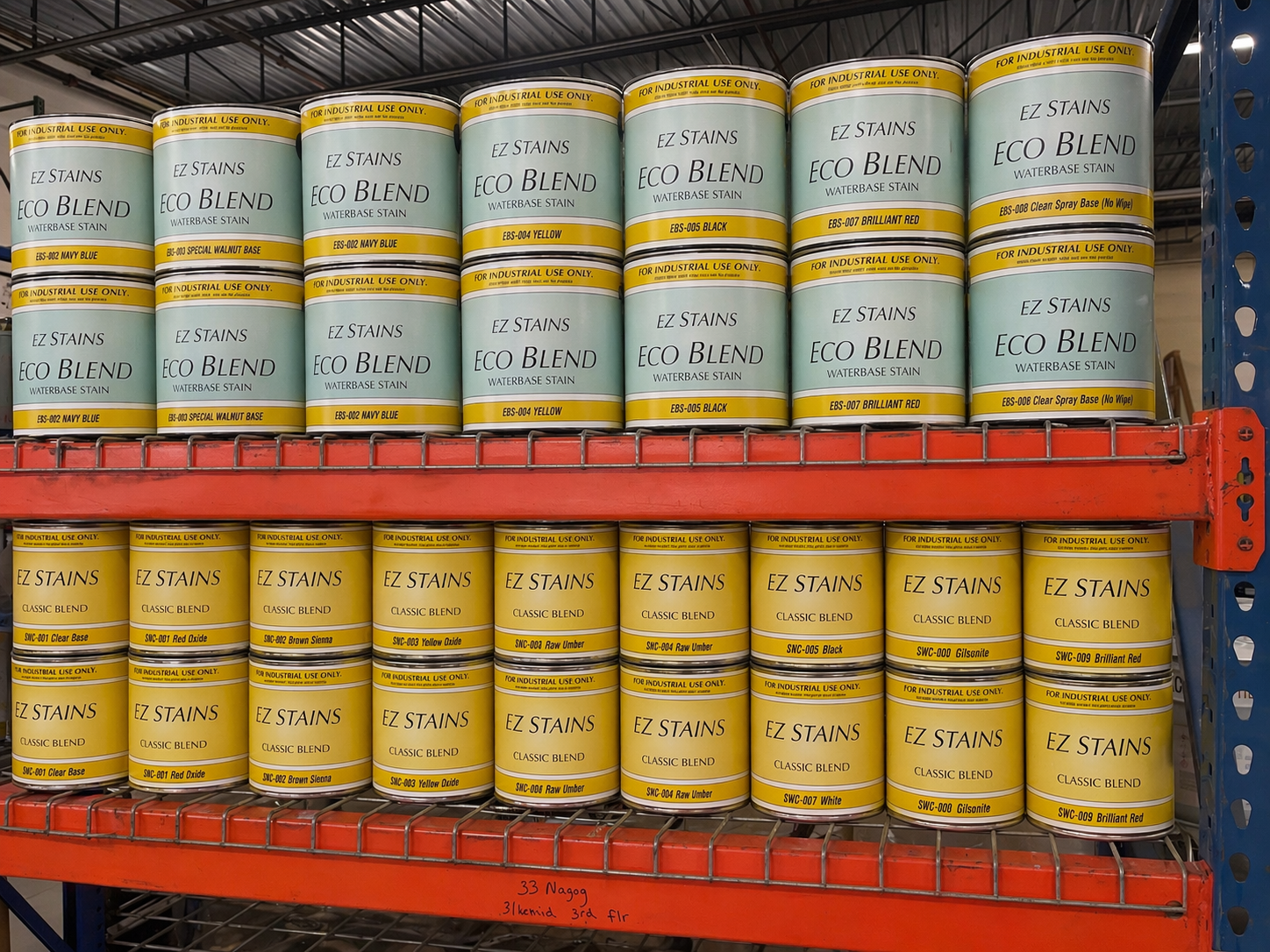 Shelves stocked with cans of EZ Stains water-based stain. The top shelf contains cans labeled with various colors including navy blue, yellow, black, and red. The bottom shelf displays cans of classic blend stain in shades like clear base, red oxide,