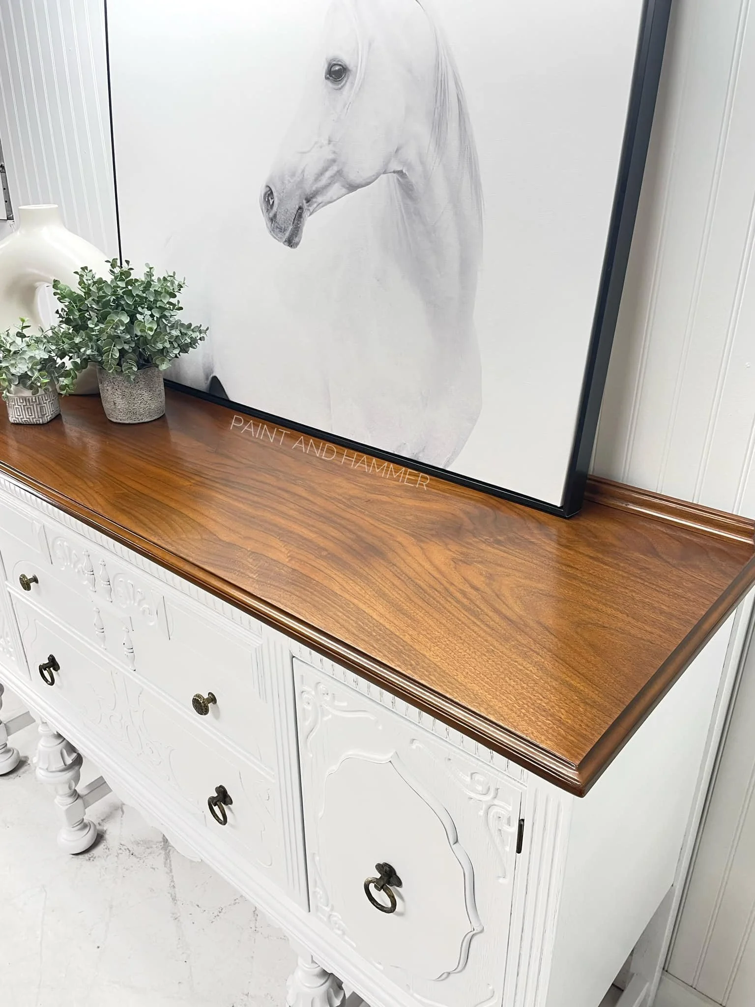 A white vintage-style sideboard with ornate details, four drawers with dark metal ring handles, and turned legs, topped with a wooden surface. On top, there are two small potted plants and a white ceramic vase.