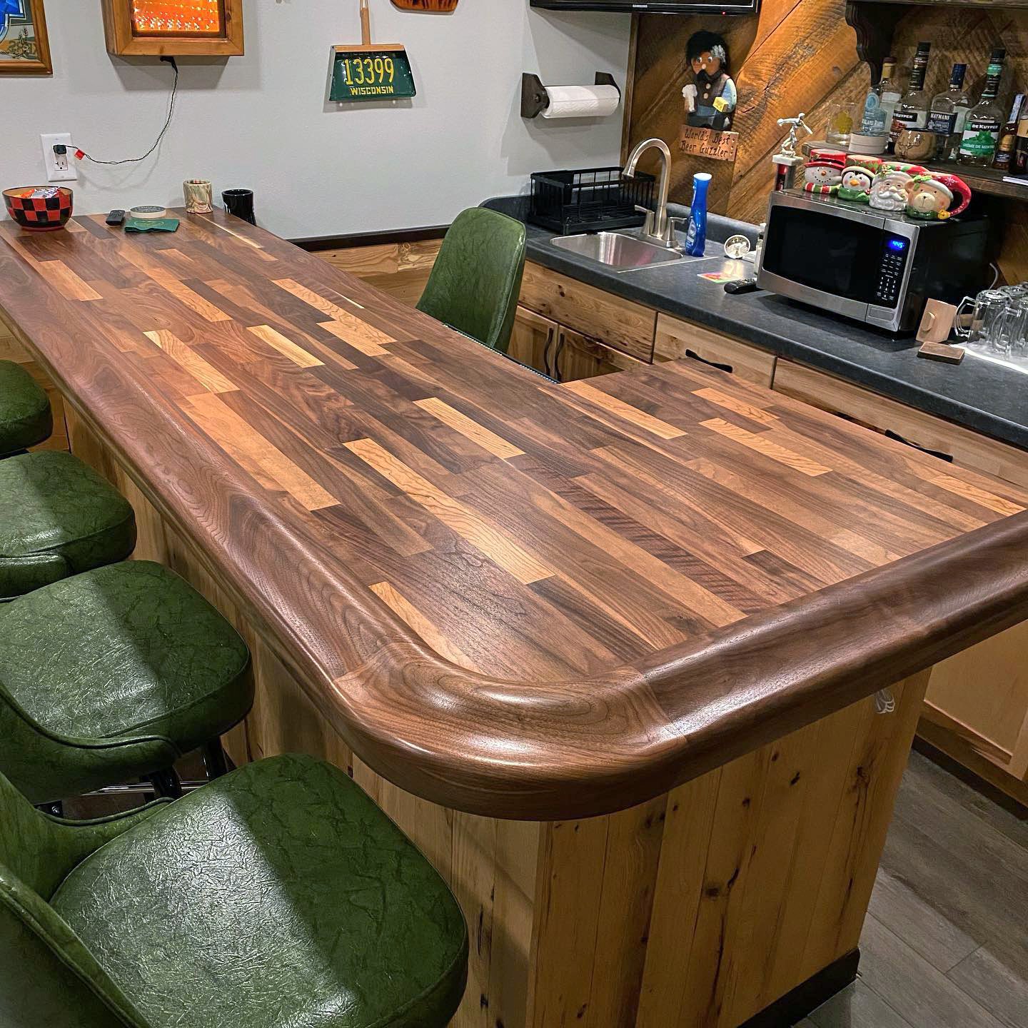A large wooden kitchen or bar counter with a polished wood surface and green leather stools around it. In the background, a sink, microwave, and various bottles and decorations are visible.