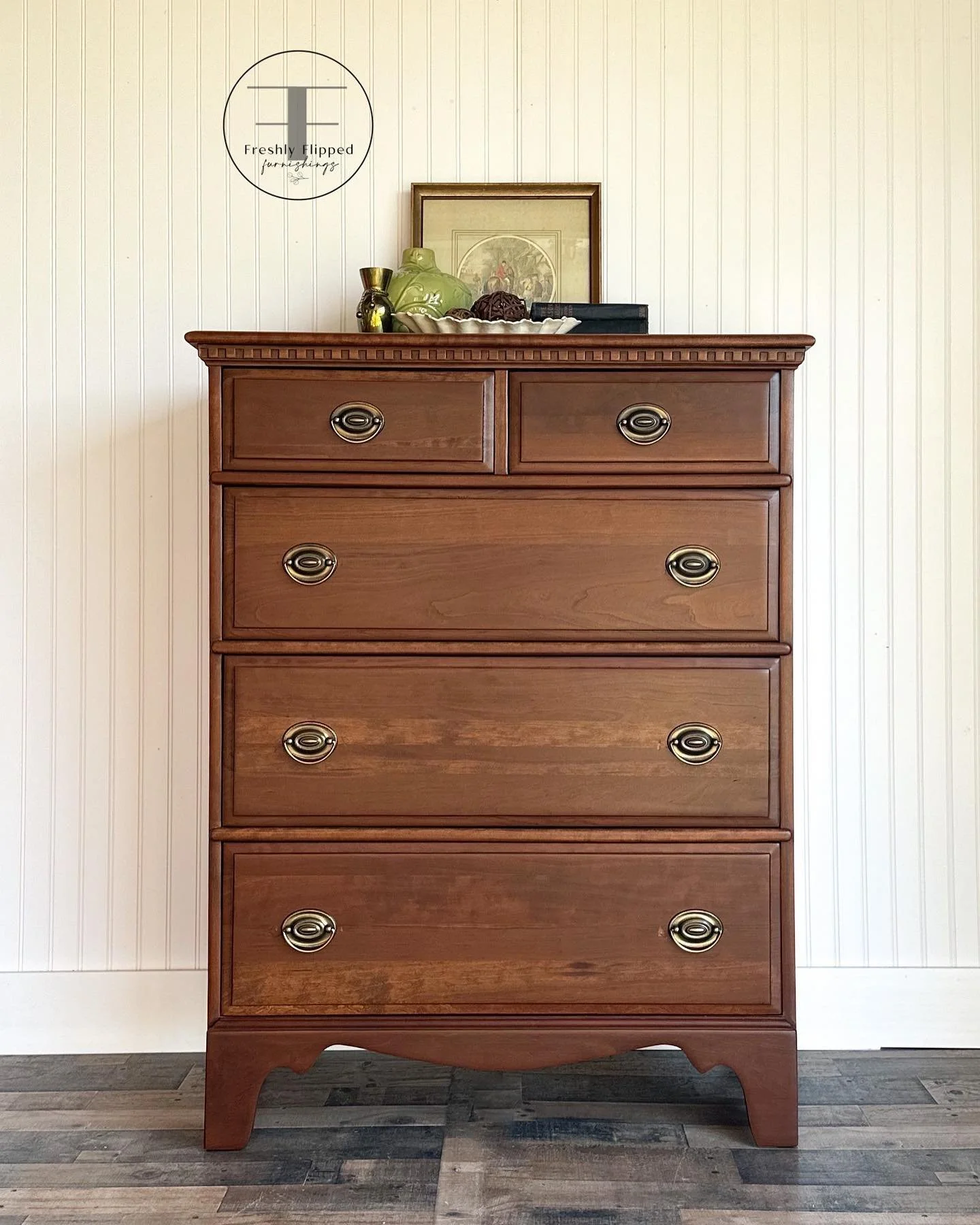 A wooden dresser with five drawers and oval metal handles, topped with decorative items and framed art, against a white paneled wall.
