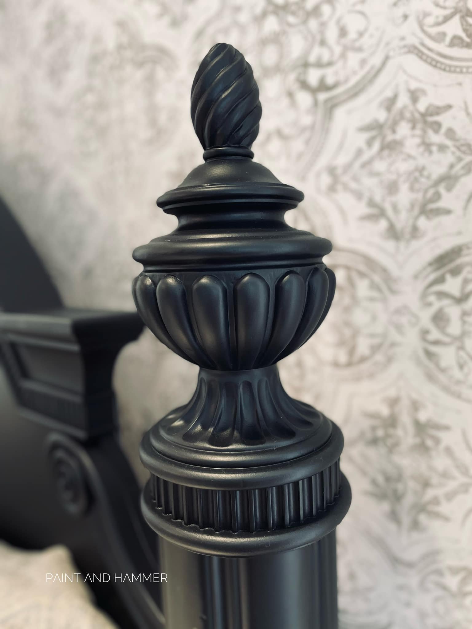 Close-up of a black decorative metal bedpost with ornate design, set against a background of patterned wallpaper.