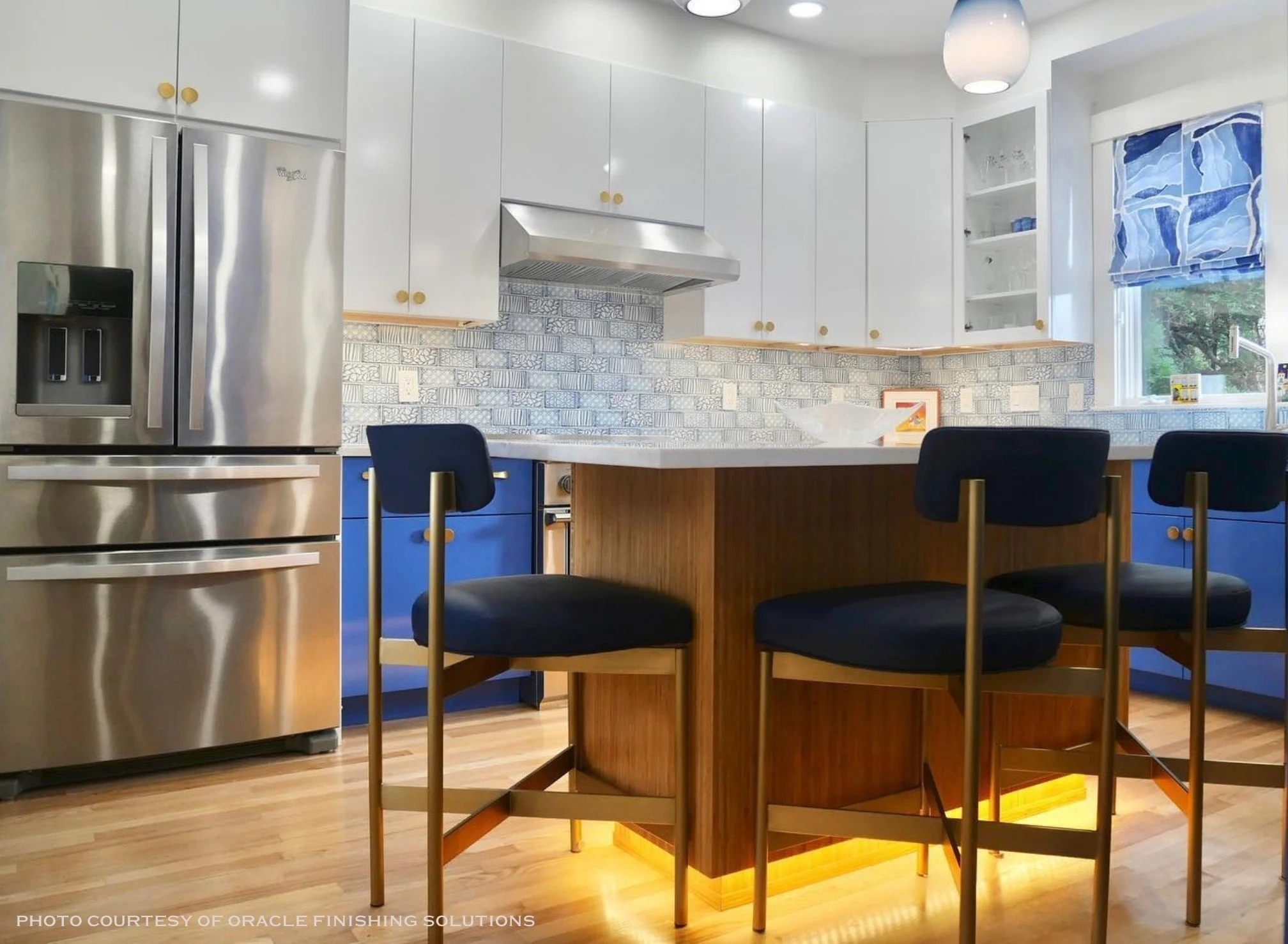 Modern kitchen with white upper cabinets, blue lower cabinets, a large stainless steel refrigerator, a wooden kitchen island with black chairs, a patterned tile backsplash, and a window with Roman shade.