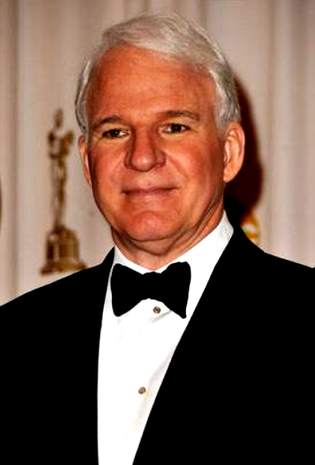 A man with white hair wearing a black tuxedo and black bow tie, standing indoors with curtains and a statue in the background.
