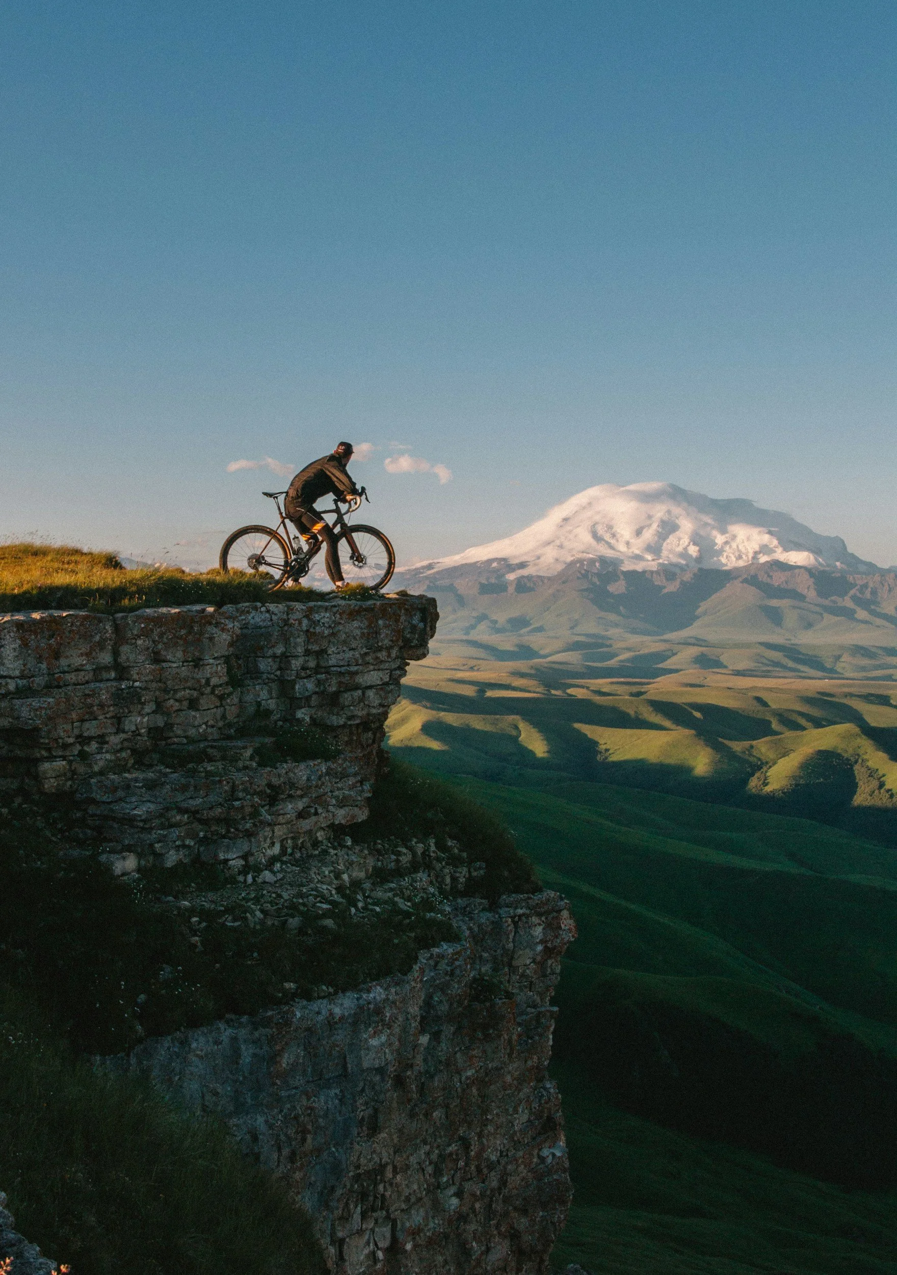 Un cycliste se tient sur le bord d'une falaise rocheuse avec un paysage de montagnes vertes et un sommet enneigé en arrière-plan, sous un ciel bleu clair.