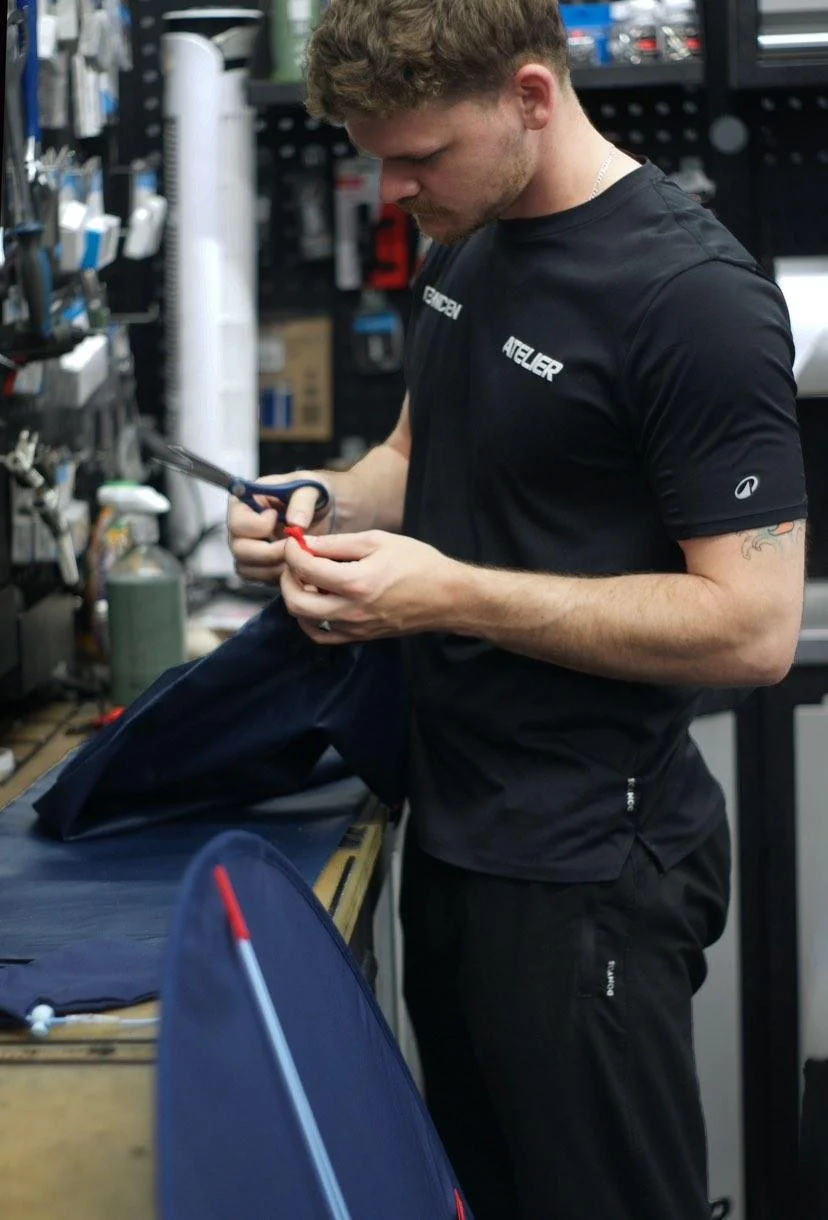 Un homme travaille dans un atelier ou un magasin, portant un t-shirt noir avec l'inscription "ENGBEN" et "ATELIER", utilisant une paire de ciseaux et des outils pour réparer ou assembler un objet en tissu ou en plastique, sur un plan de travail en bois.