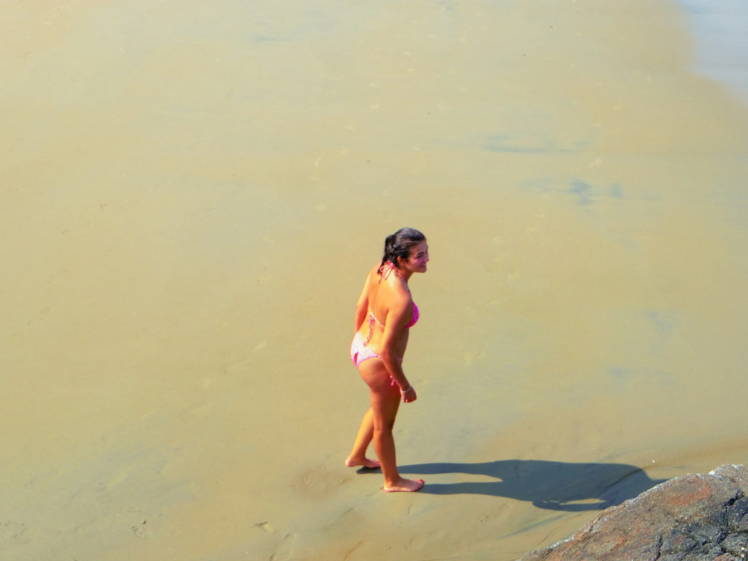 A woman in a pink bikini standing at the edge of a sandy beach, looking towards the water with her shadow cast on the sand.