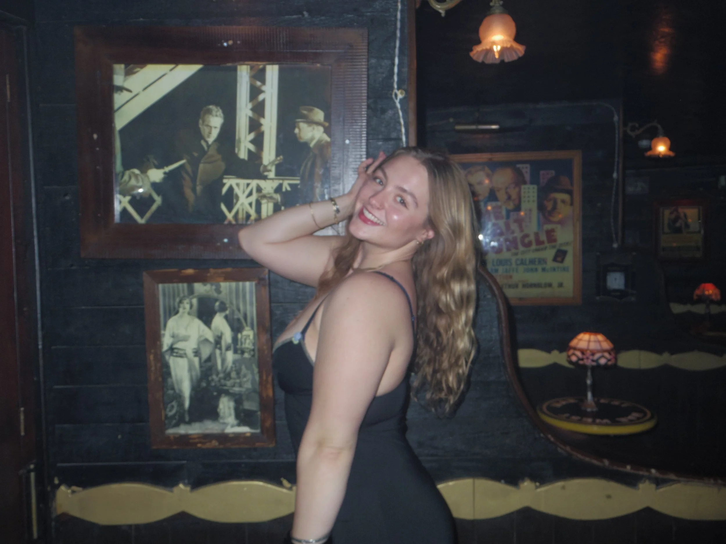 A smiling woman with wavy blonde hair posing inside a dimly lit room decorated with vintage posters and framed photographs on the dark wall. She is touching her hair and looking at the camera.