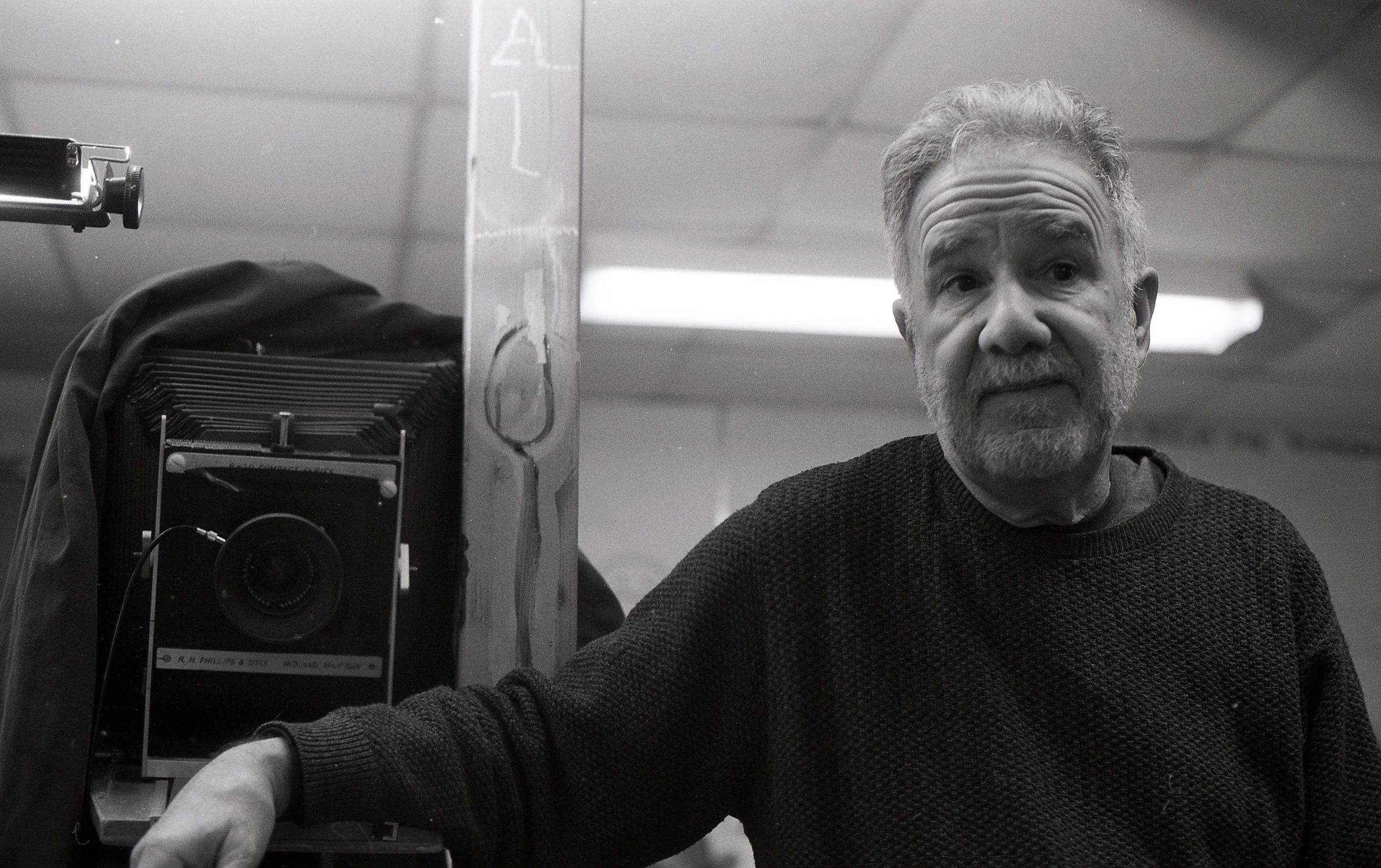 A black-and-white photo of an older man with gray hair and a beard, standing behind a camera on a tripod, in what appears to be a photo studio or darkroom. The man is wearing a dark sweater and looking at the camera.