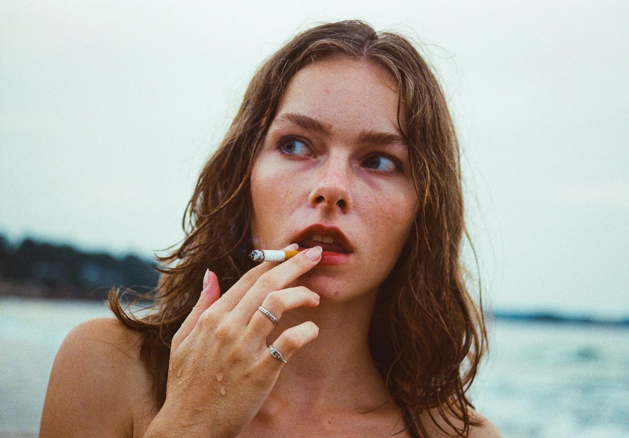 A young woman with wet, wavy brown hair holding a cigarette near her lips, outdoors with blurred background.