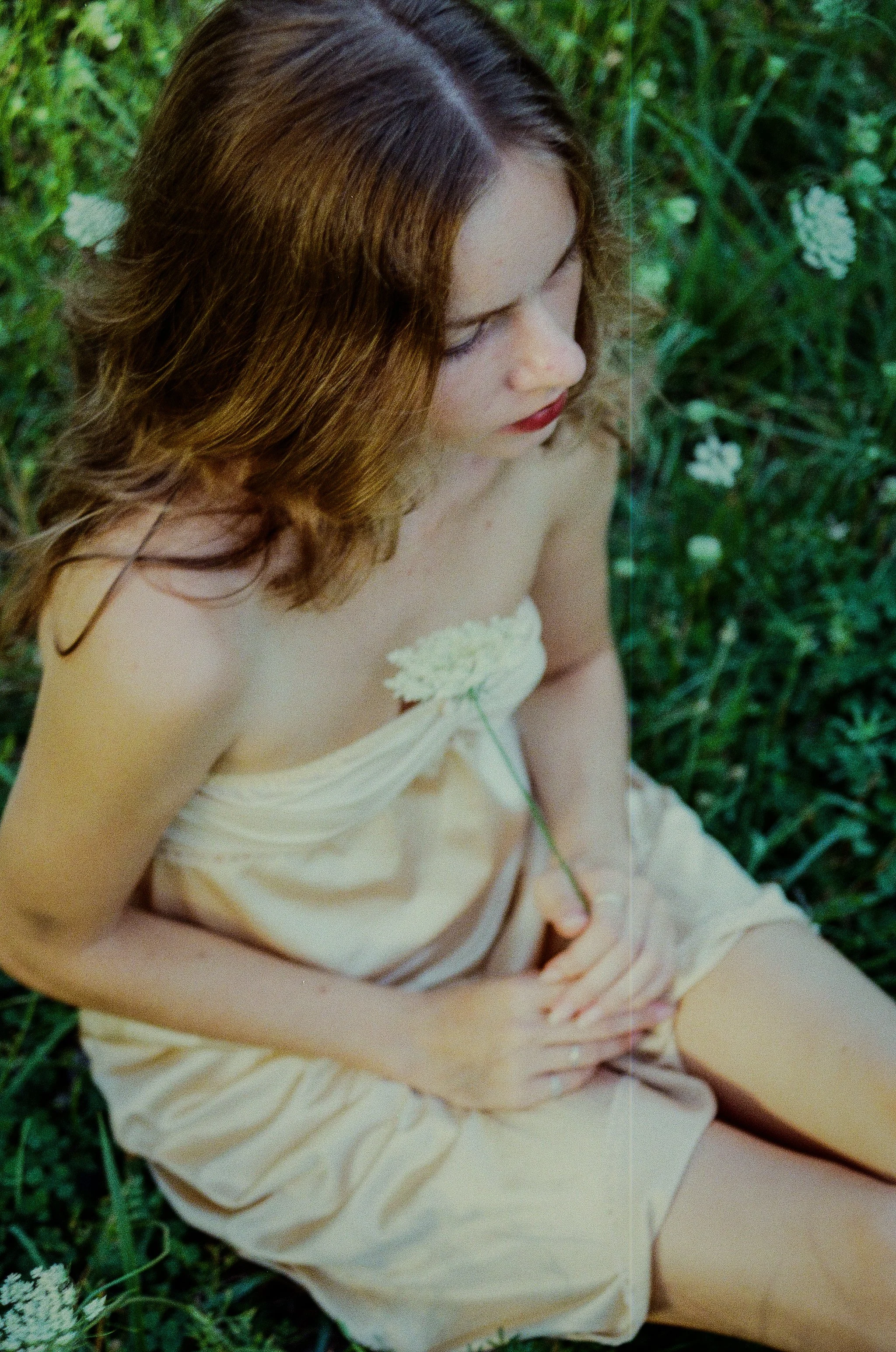 A woman with long wavy brown hair, wearing a light-colored dress, sitting in a green grassy field.