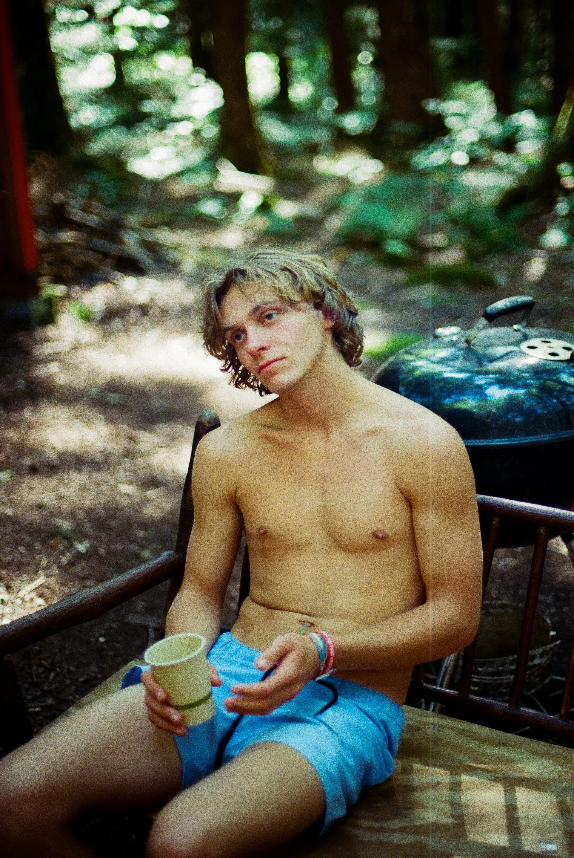 A shirtless young man with curly blond hair sitting outdoors in a wooded area, holding a white cup in his right hand, with a black helmet and a coffee pot or thermos behind him.