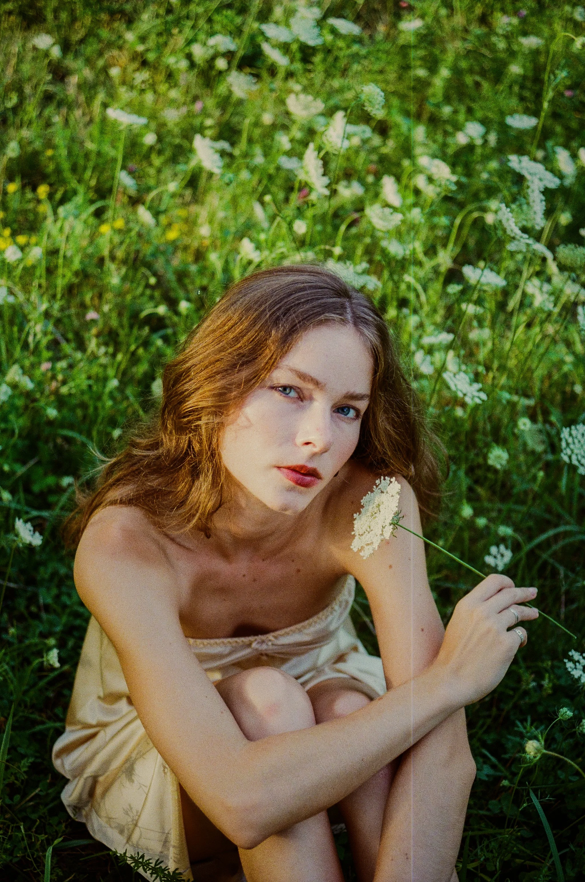 A young woman with wavy auburn hair and blue eyes sitting in a green field of wildflowers, holding a flower stem in her hand, dressed in a light-colored dress.