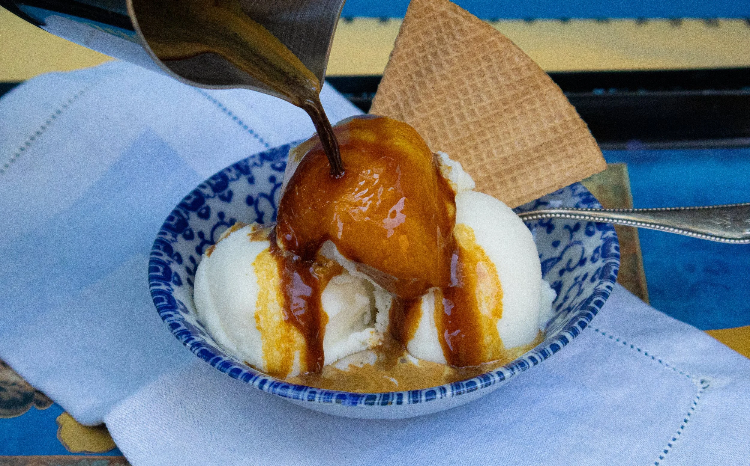 Vanilla ice cream topped with caramel sauce and a whole caramelized pear, served in a patterned blue and white bowl with a wafer and a spoon.