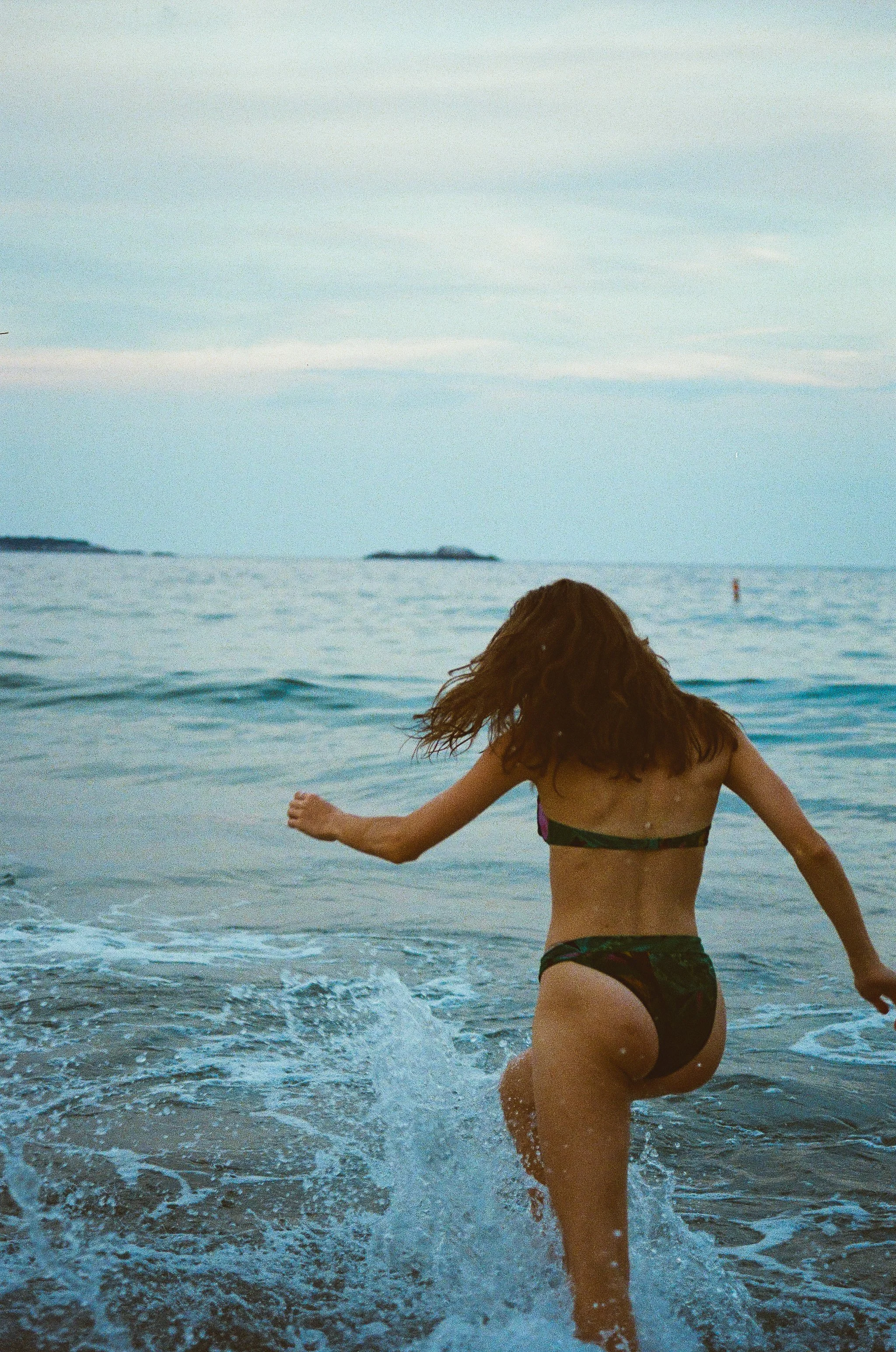 A woman with curly hair in a bikini running into the ocean at the beach during daytime, with boats and islands visible in the distance.