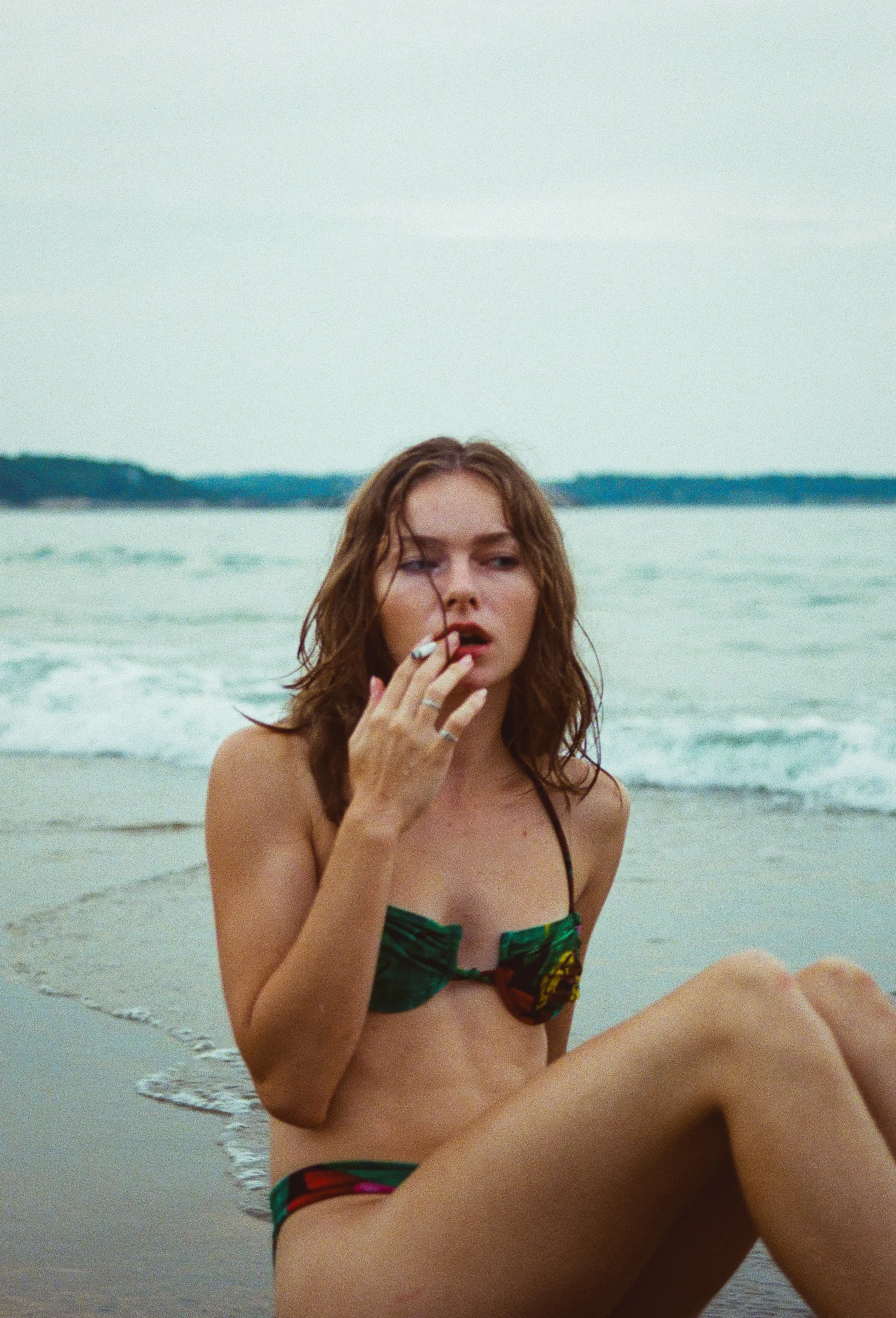 A young woman with wet hair sitting on the beach in a colorful bikini, holding a cigarette near her mouth, with the ocean and sky in the background.