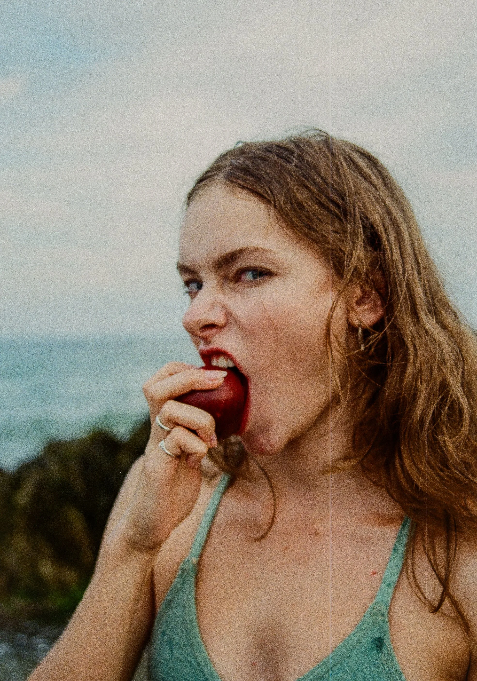 A young woman with red hair and blue eyes is eating a red apple outdoors near water, wearing a green spaghetti strap top.