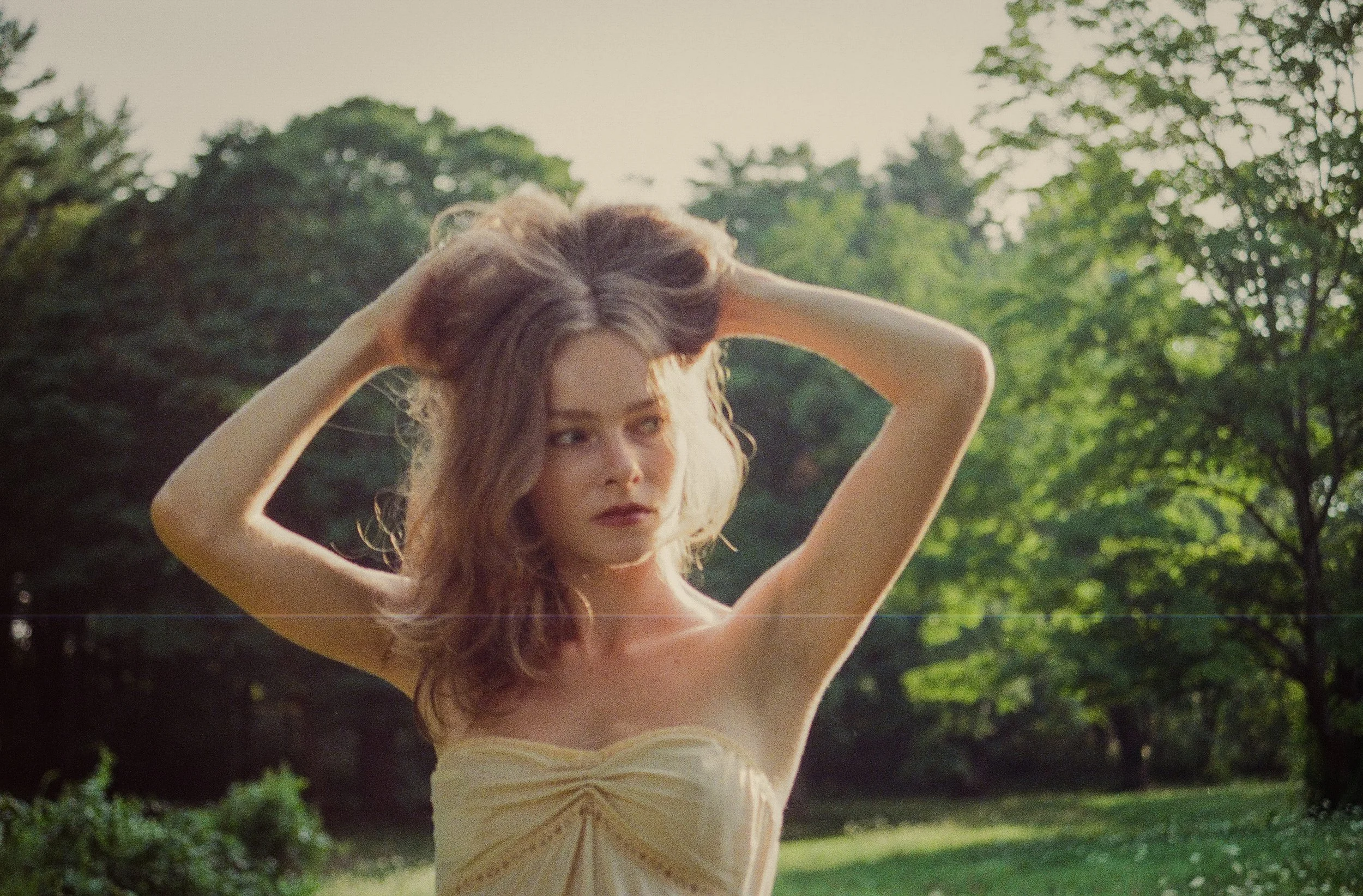 A woman with curly hair standing outdoors in a park, holding her hair back with her hands, wearing a strapless dress, with trees and greenery in the background.