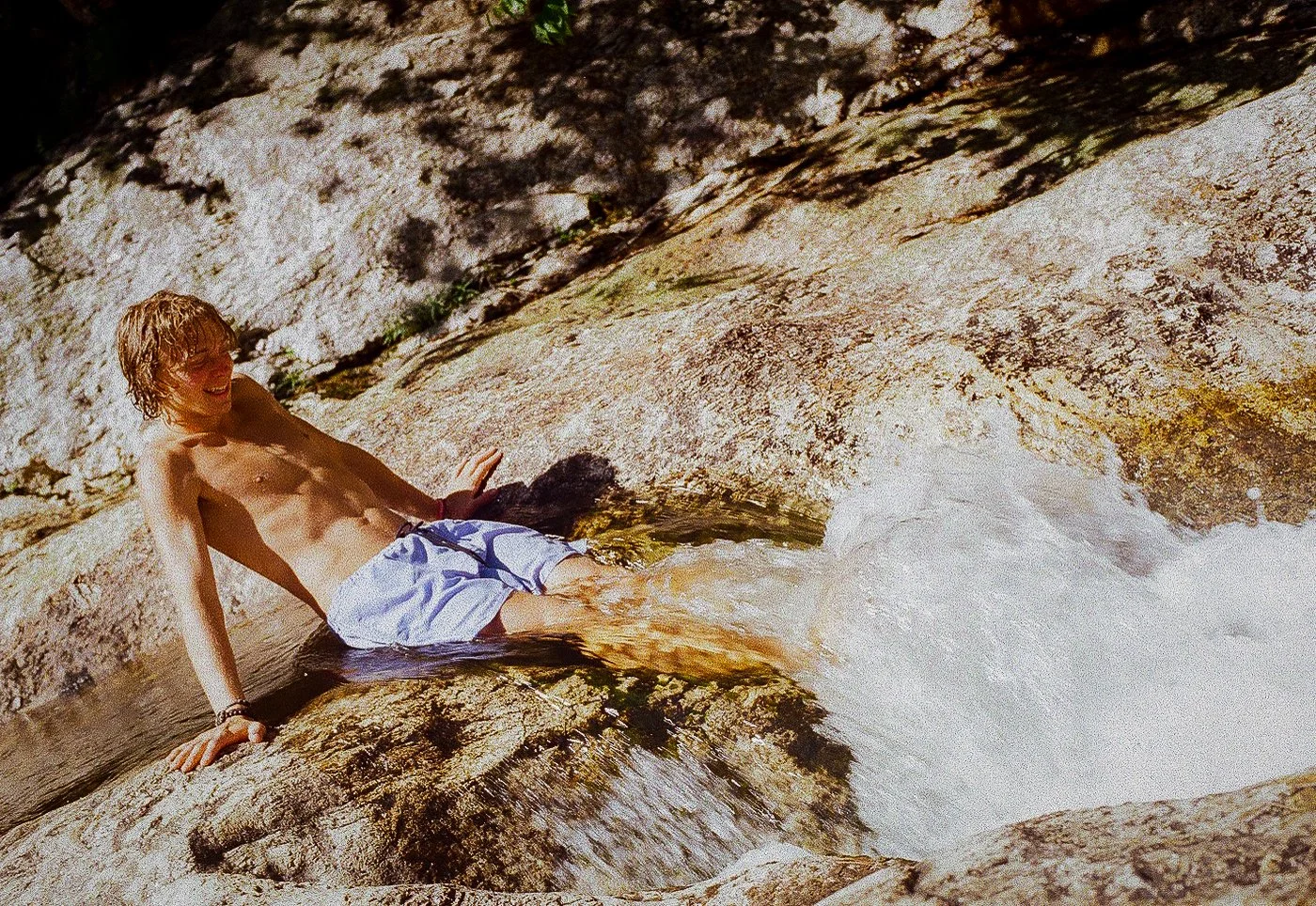A young person with wet hair sitting on rocks next to a rushing stream of water, smiling.