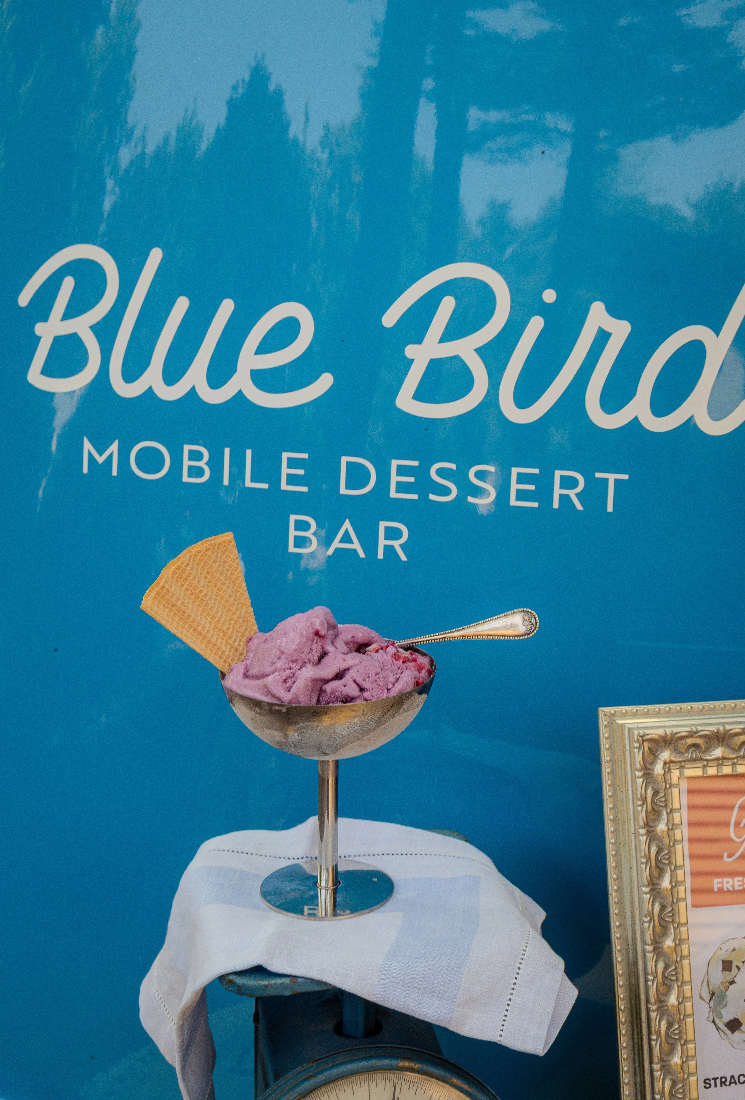 A sign for Blue Bird Mobile Dessert Bar featuring a bowl of purple ice cream with a waffle cone and a spoon, set against a blue sky background.