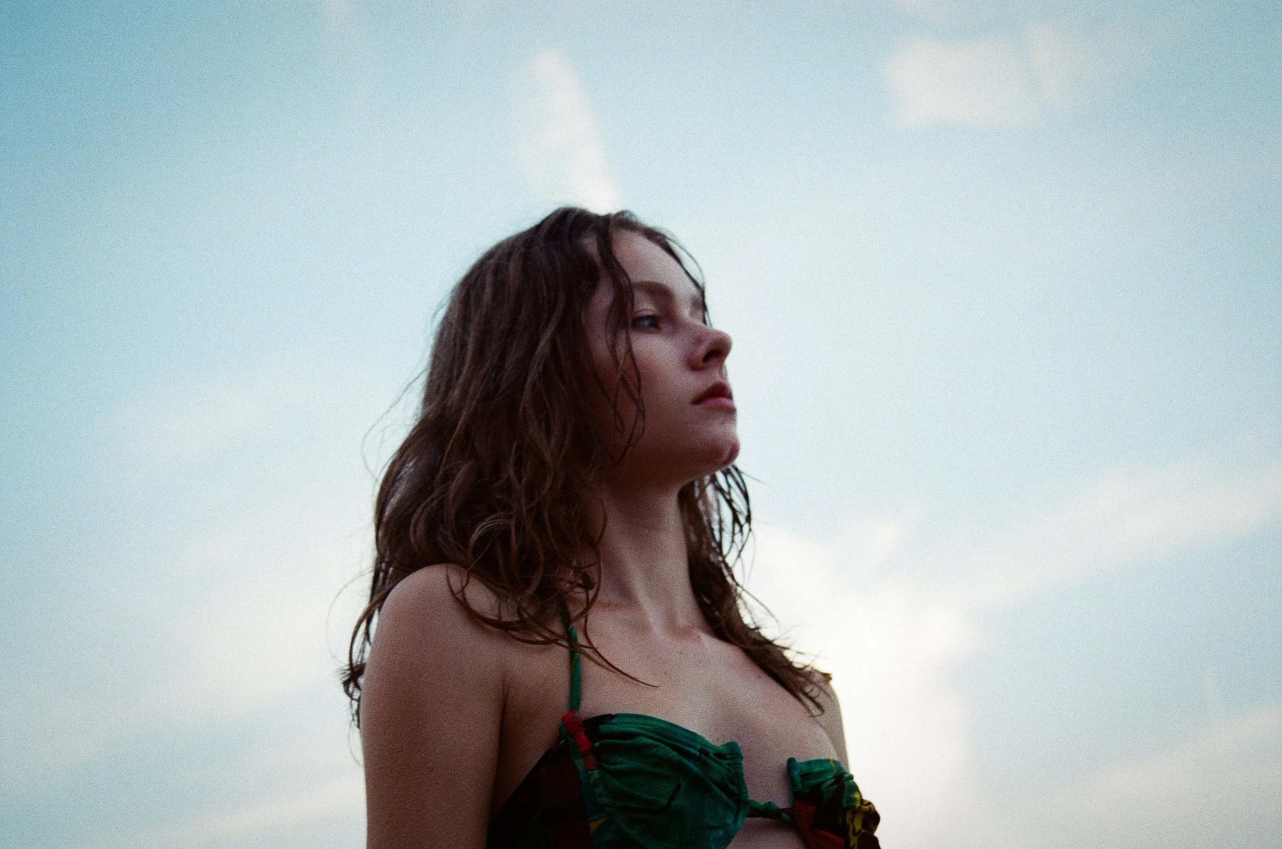 A woman with wet, curly brown hair looking into the distance against a cloudy sky. She is wearing a green and black patterned top.