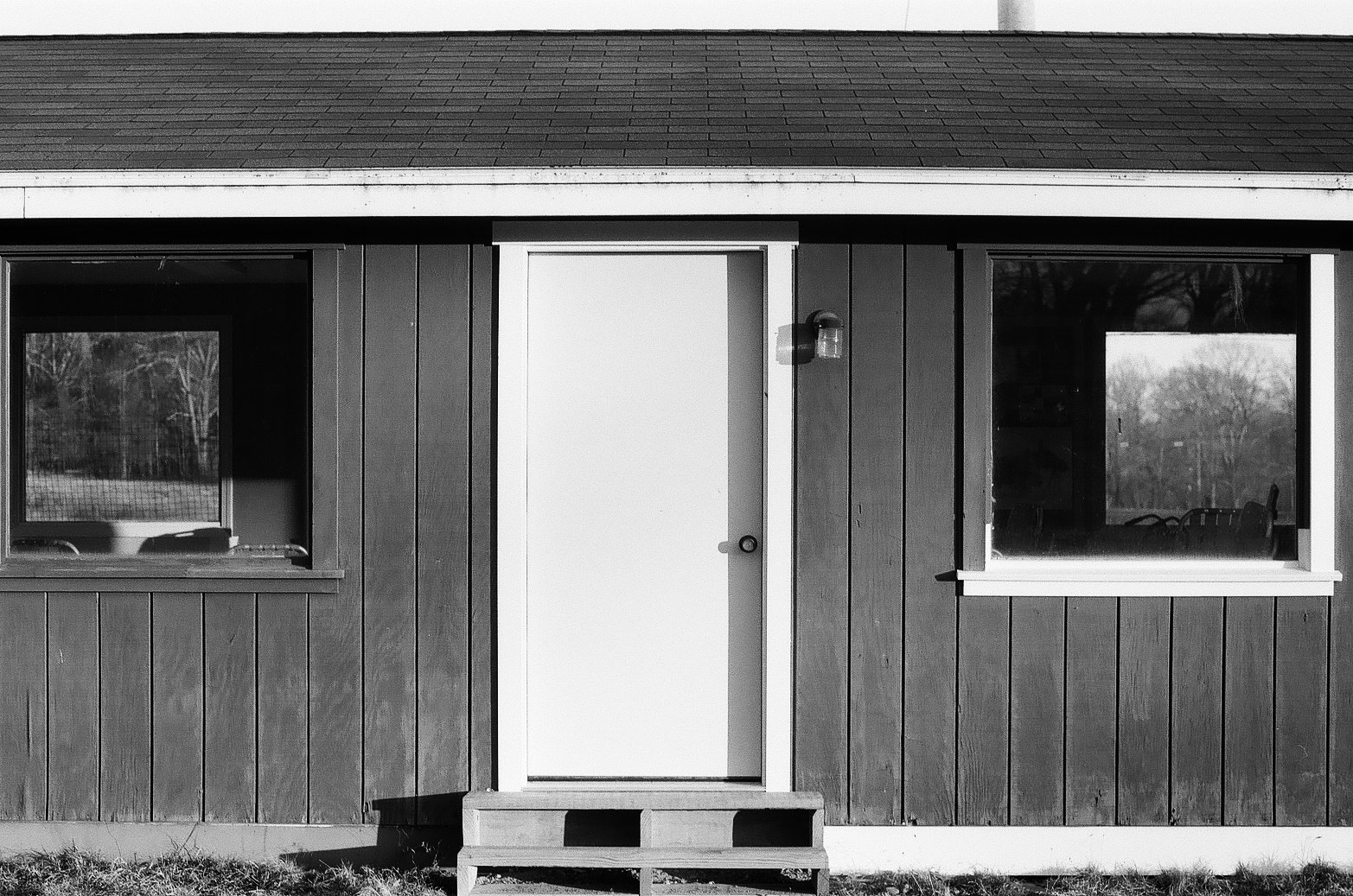 Front view of a small house with wooden exterior, two rectangular windows, white door with a small step, and exterior light fixture.