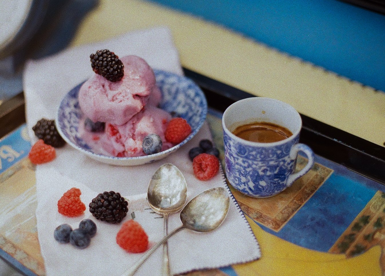 A bowl of pink berry sorbet with blackberries, blueberries, and raspberries on top, accompanied by a cup of coffee and two silver spoons, all on a white napkin with scattered berries.
