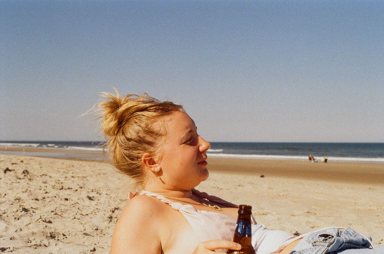 A woman with blonde hair in a messy bun relaxing on a sandy beach, holding a beer, with ocean waves in the background.