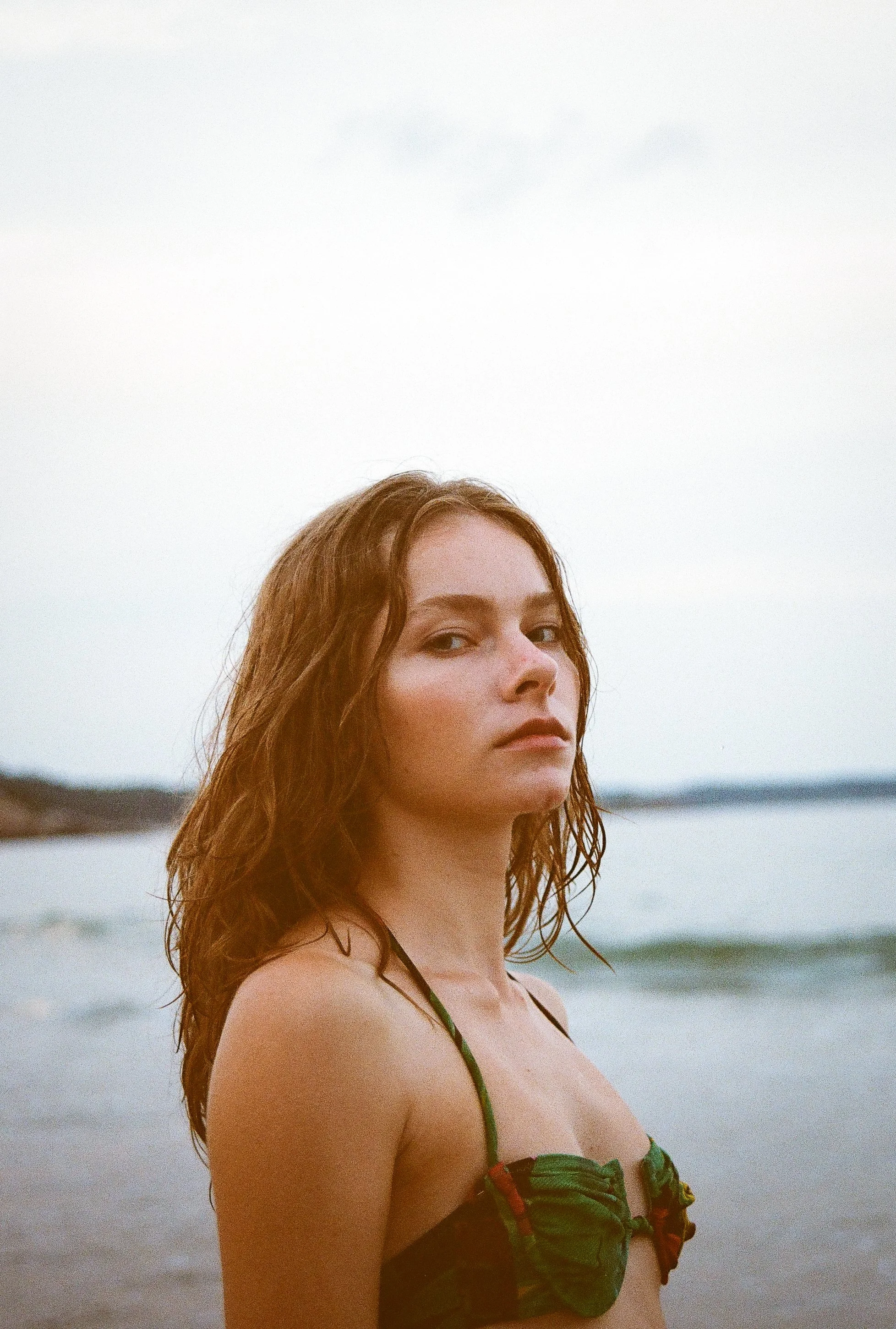 A woman with red hair wearing a bikini top standing on a beach with water and distant shoreline in the background