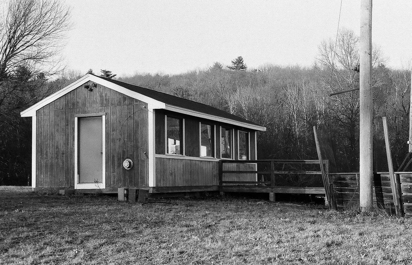 Black and white photo of a small wooden house with a sloped roof, a door on the front, and a deck with a railing. There is a utility pole with wires on the right, and leafless trees in the background.
