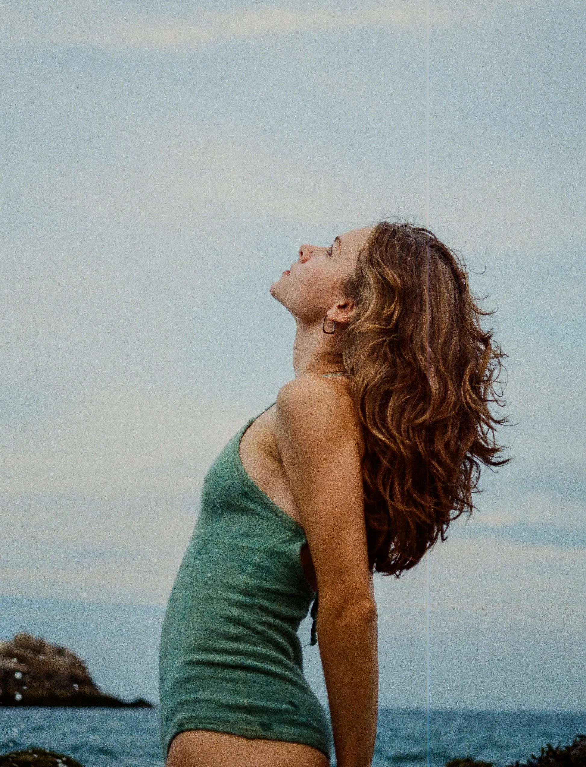 A woman with curly hair standing outdoors near the water, looking up with her eyes closed, in a scenic coastal area.