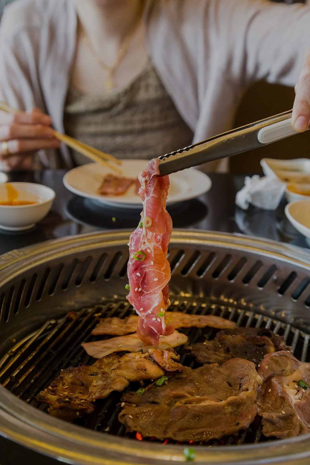 Customer using tongs to place fresh, premium cuts of raw meat onto a hot table-side grill for an authentic Korean BBQ experience in Brentwood, Tennessee.