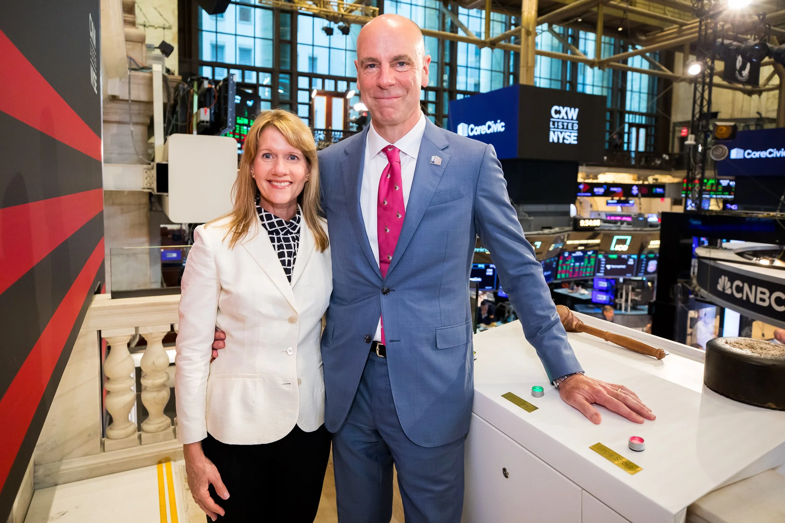 A woman and a man in business attire standing together on a trading floor, with stock market screens and logos in the background.
