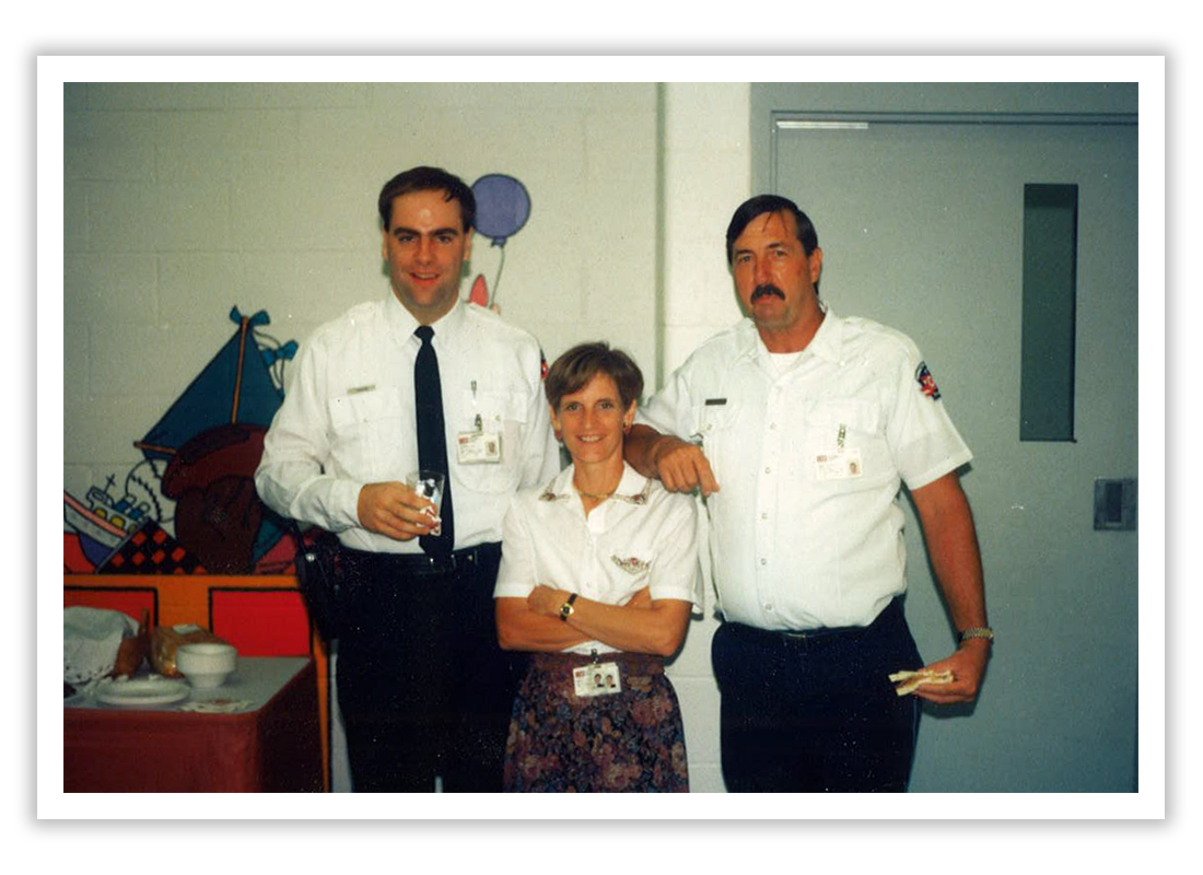 Three people in uniform, two men and one woman, standing indoors with a decorated wall in the background. They are smiling and posing for the photo.