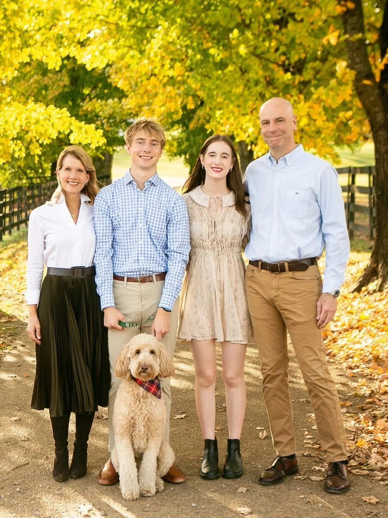 A family of five, including a golden doodle dog, standing outdoors on a pathway with autumn trees and leaves in the background, all smiling.