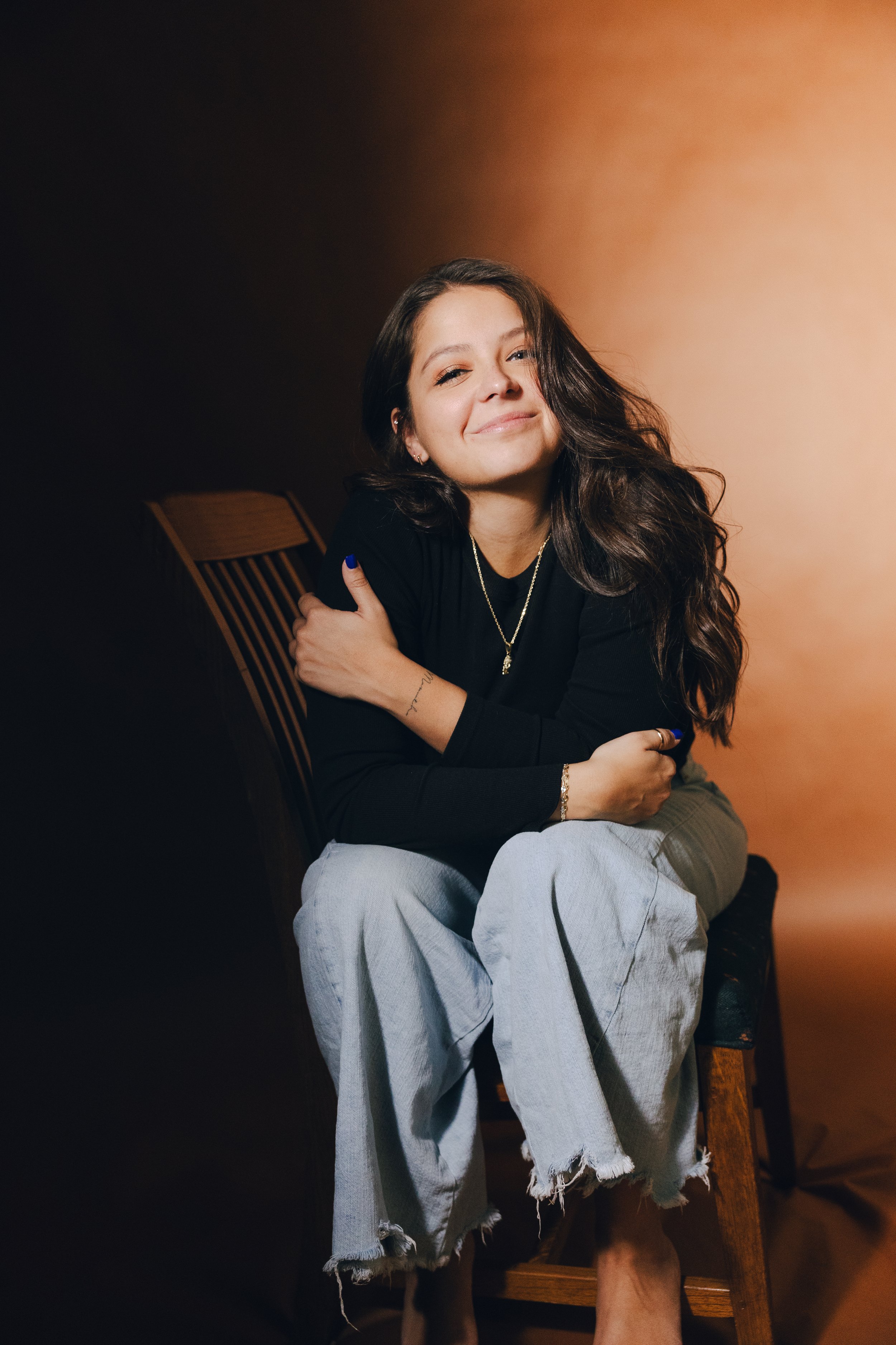 A young woman with long wavy brown hair, sitting on a wooden chair, smiling, wearing a black top, light gray ripped jeans, jewelry, and dark blue nail polish, against a gradient background from dark to light