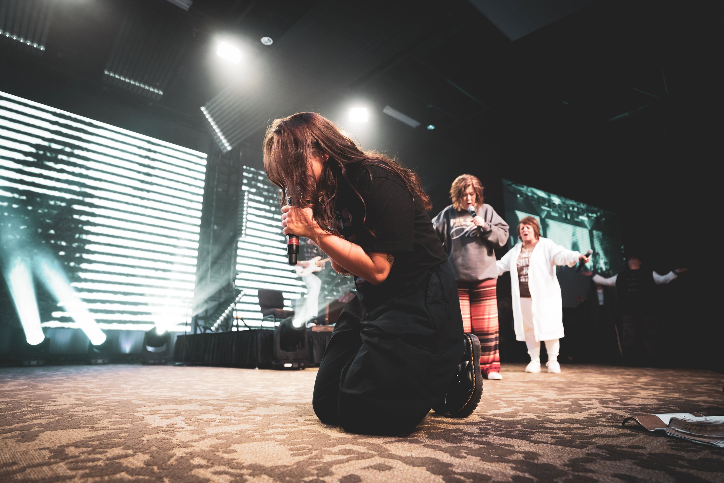 People praying and singing on stage during a concert or worship event with a large digital screen in the background.