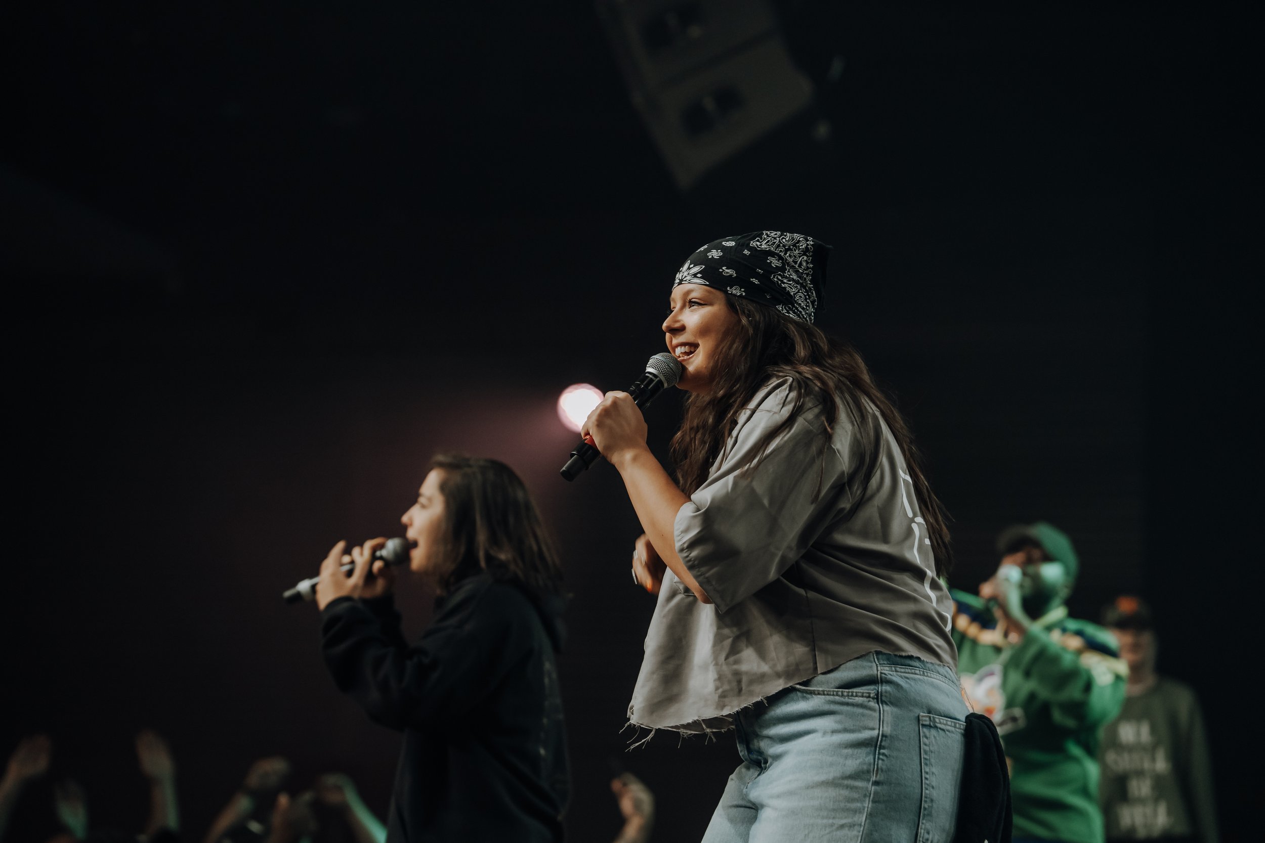 Two women singing with microphones on a dark stage, one wearing a bandana and a gray shirt, the other wearing a black jacket, while a person in green plays a flute in the background.