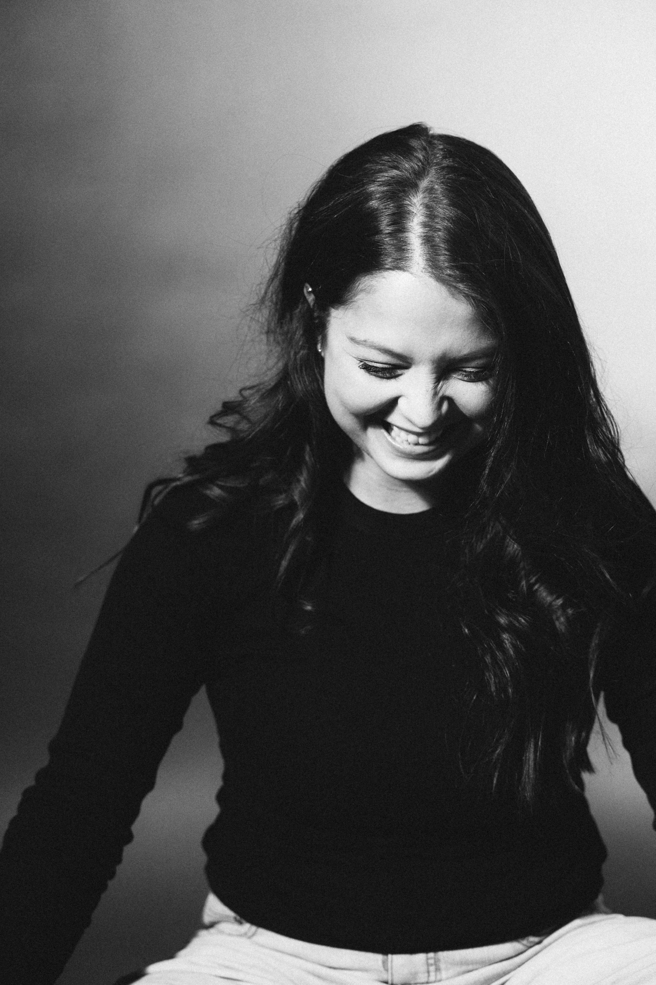 A black and white photo of a woman laughing, with long dark hair, wearing a dark top and light pants, looking downward.