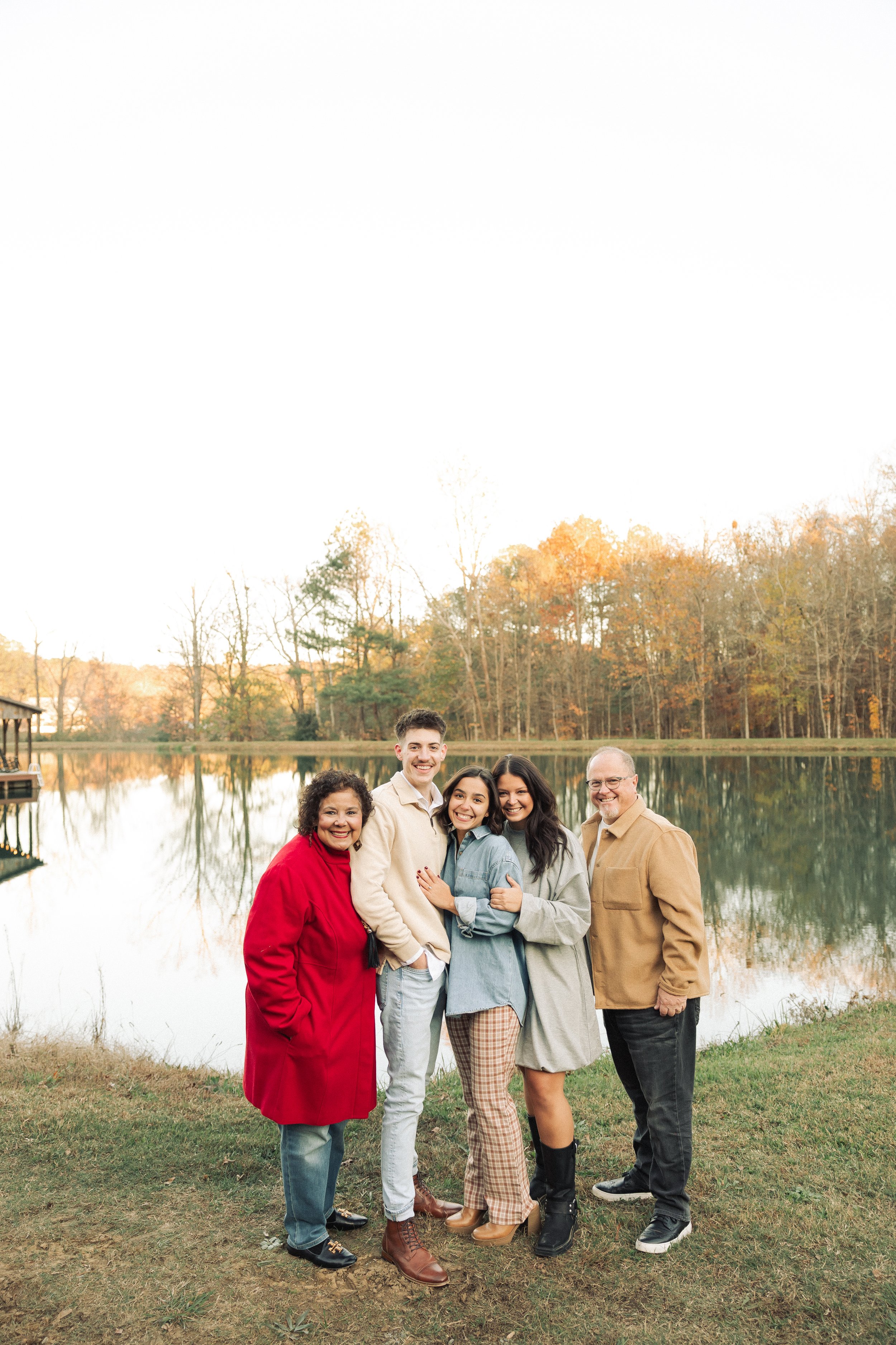 A group of five people standing by a lake, smiling and posing for a photo during autumn, with trees showing fall foliage in the background.