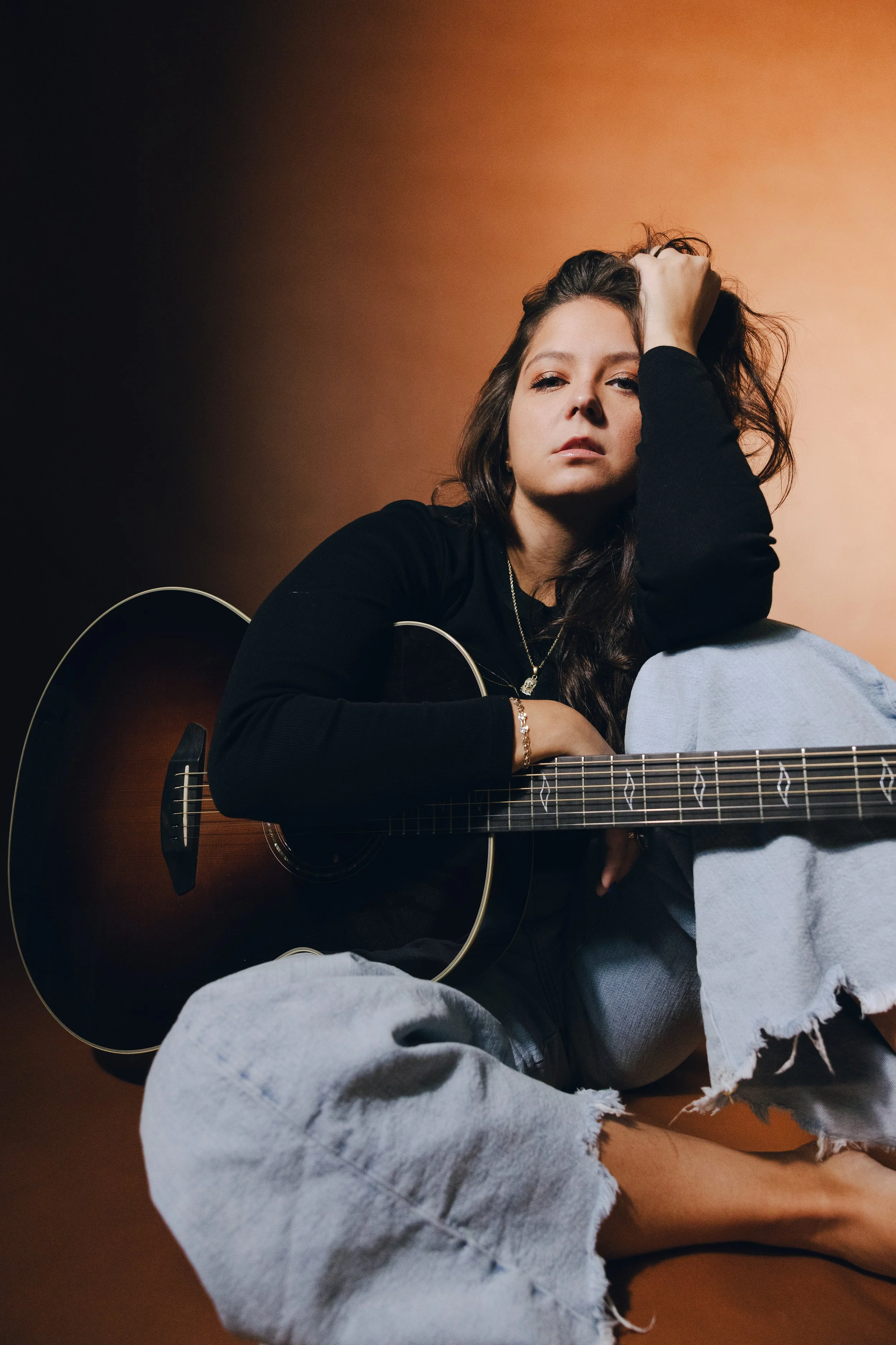 A young woman with long dark hair, dressed in a black top and ripped light blue jeans, sitting barefoot against a warm brown background, holding an acoustic guitar.