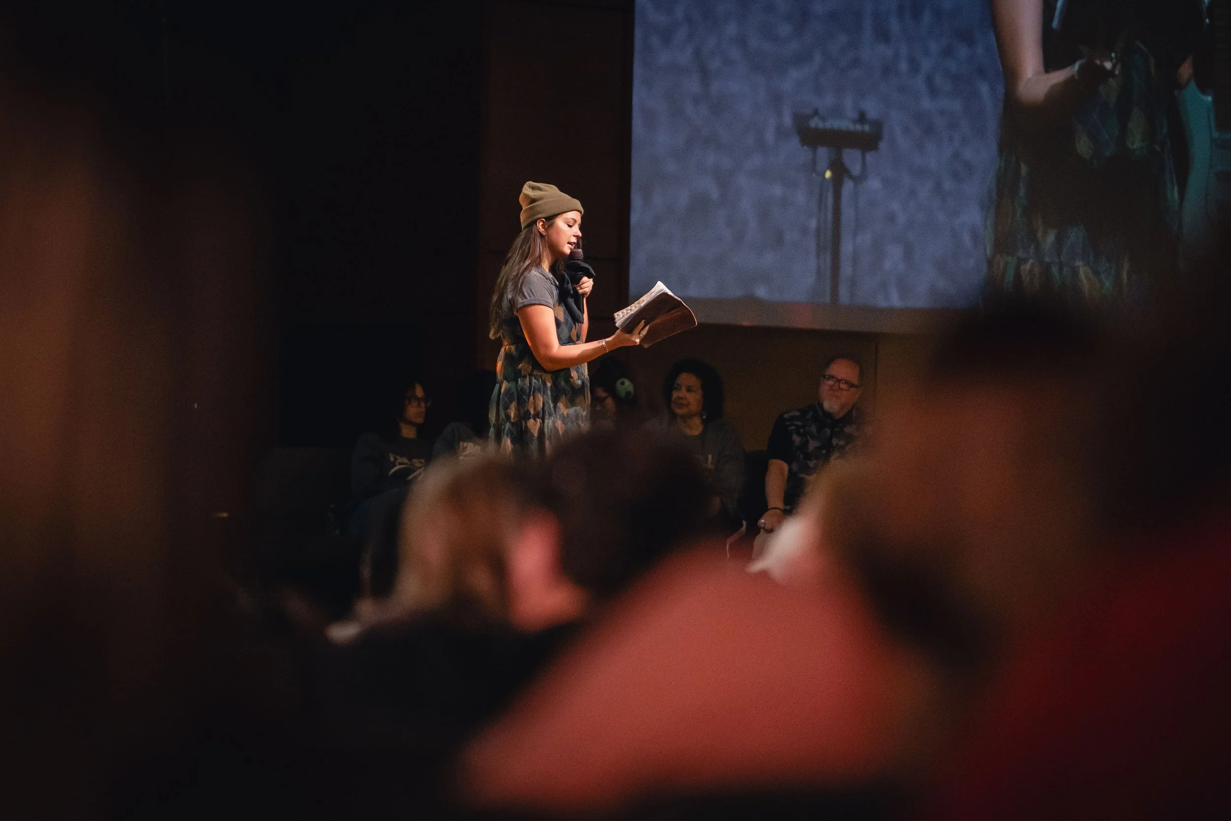 A young woman wearing a beanie hat and patterned dress reads from a book while speaking into a microphone on stage. Several people sit in the background watching her, and a large screen behind displays her image.