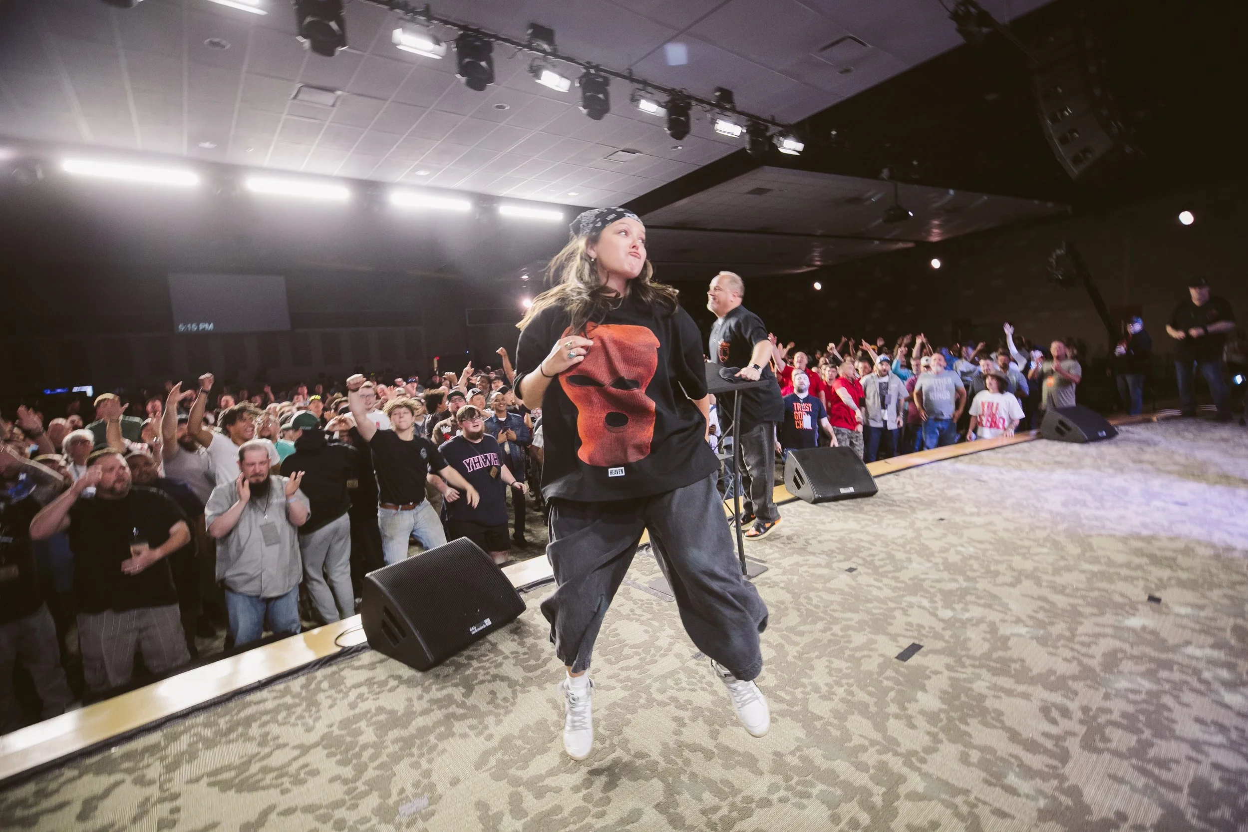 Crowd of people dancing and celebrating on stage during a concert or event in an indoor venue with stage lights overhead.