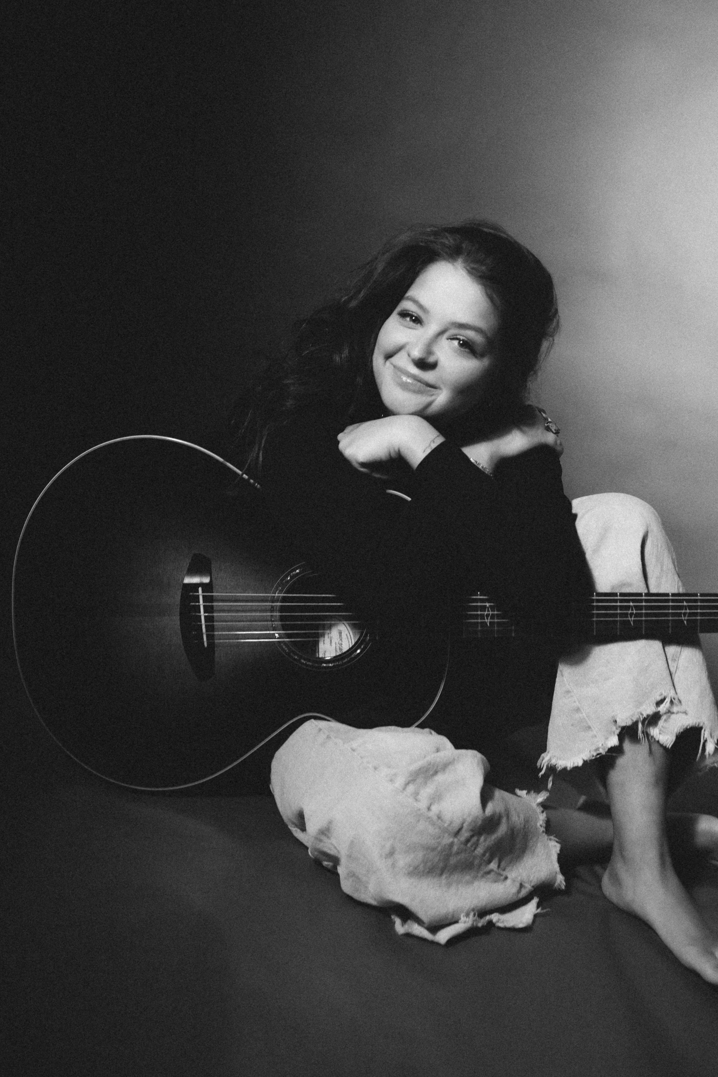 A young woman with dark wavy hair smiling, sitting with a guitar, wearing ripped jeans, in a black and white portrait.
