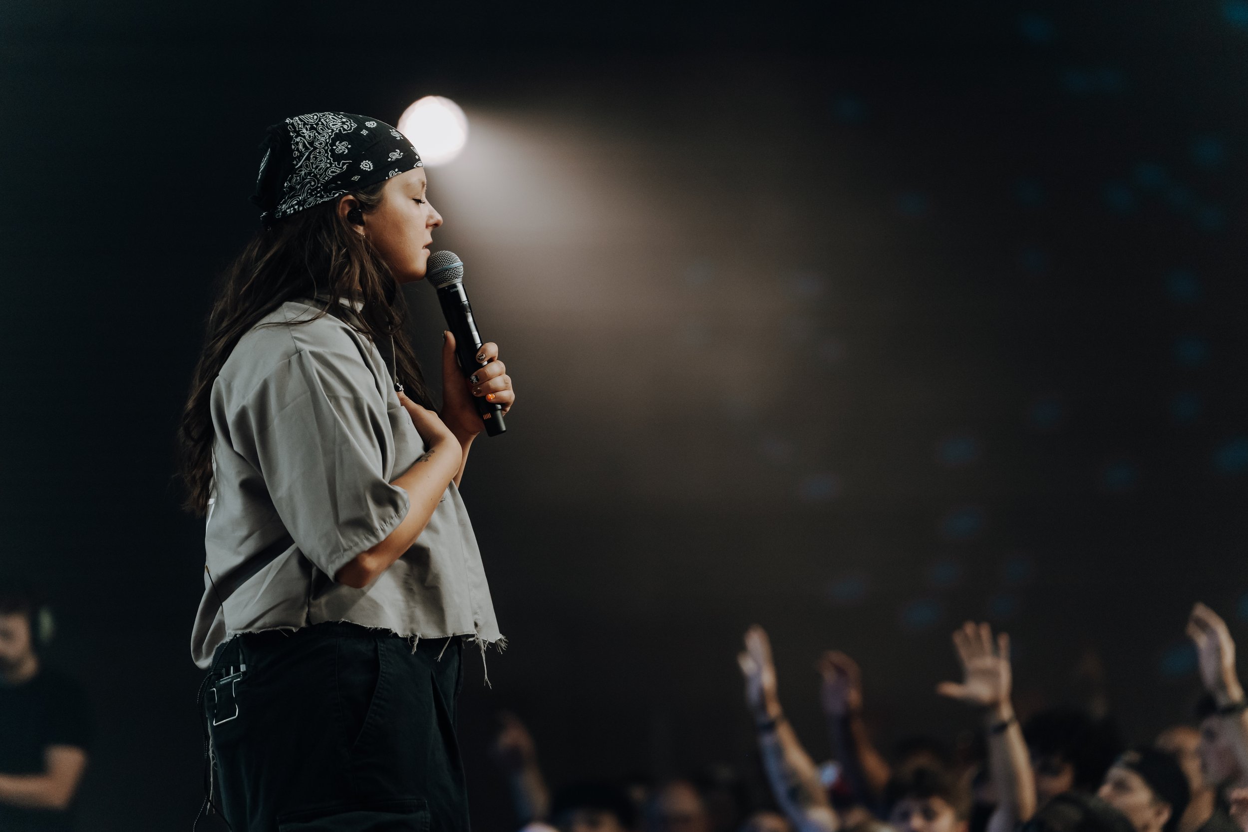A young woman with a bandana on her head singing into a microphone in front of a dark audience with raised hands.