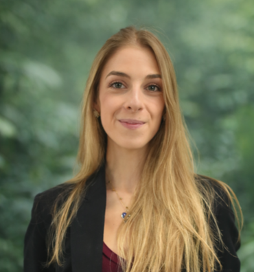Young woman with long blonde hair wearing a black blazer and maroon top, standing outdoors with green trees in the background.