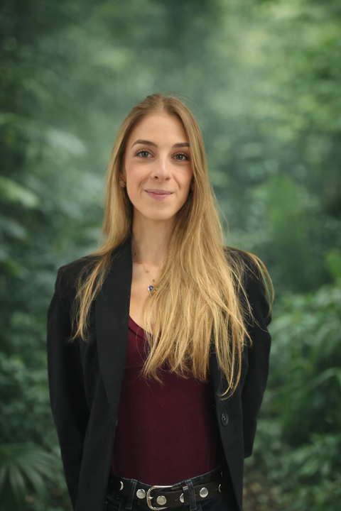 Young woman with long blonde hair wearing a black blazer and maroon top, standing outdoors with green trees in the background.