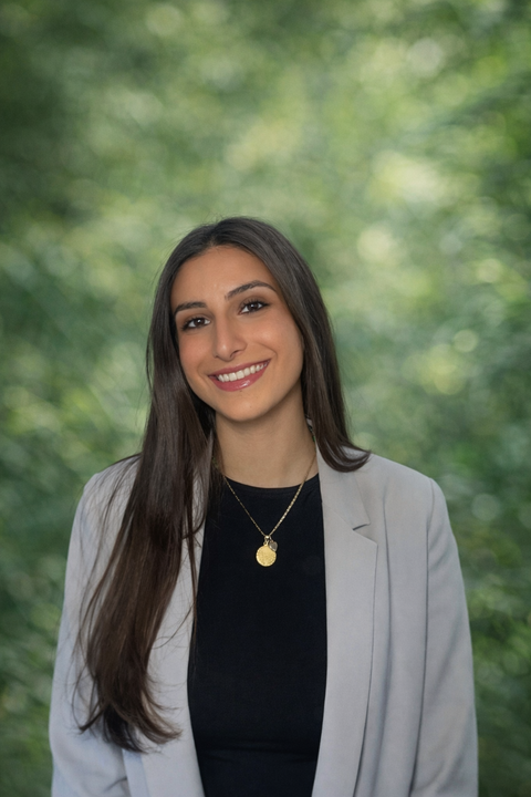 A young woman with long dark hair, smiling, wearing a gray blazer over a black top, with a gold necklace, standing outdoors with blurred green trees in the background.