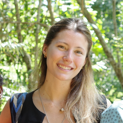A smiling young woman with long, light brown hair standing outdoors in a green, wooded area.