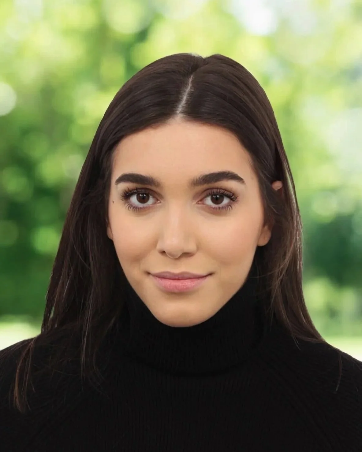A woman with dark brown hair and brown eyes wearing a black turtleneck, standing against a blurred green outdoor background.